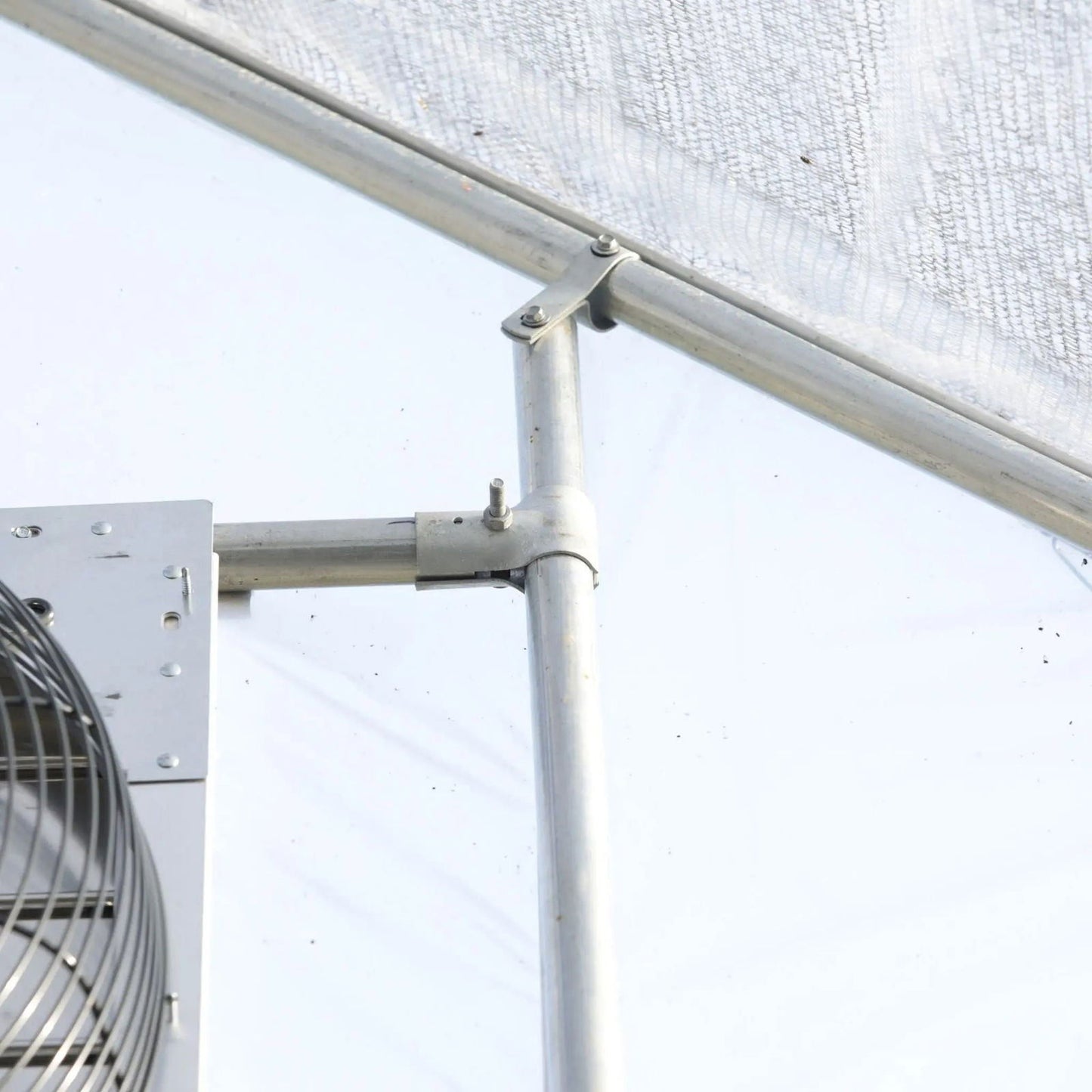 A close-up of Bootstrap Farmer's Greenhouse Shutter Mount Exhaust Fans shows the metal pipe joint and support bracket attached to a hoop house frame, with translucent greenhouse panels in the background.