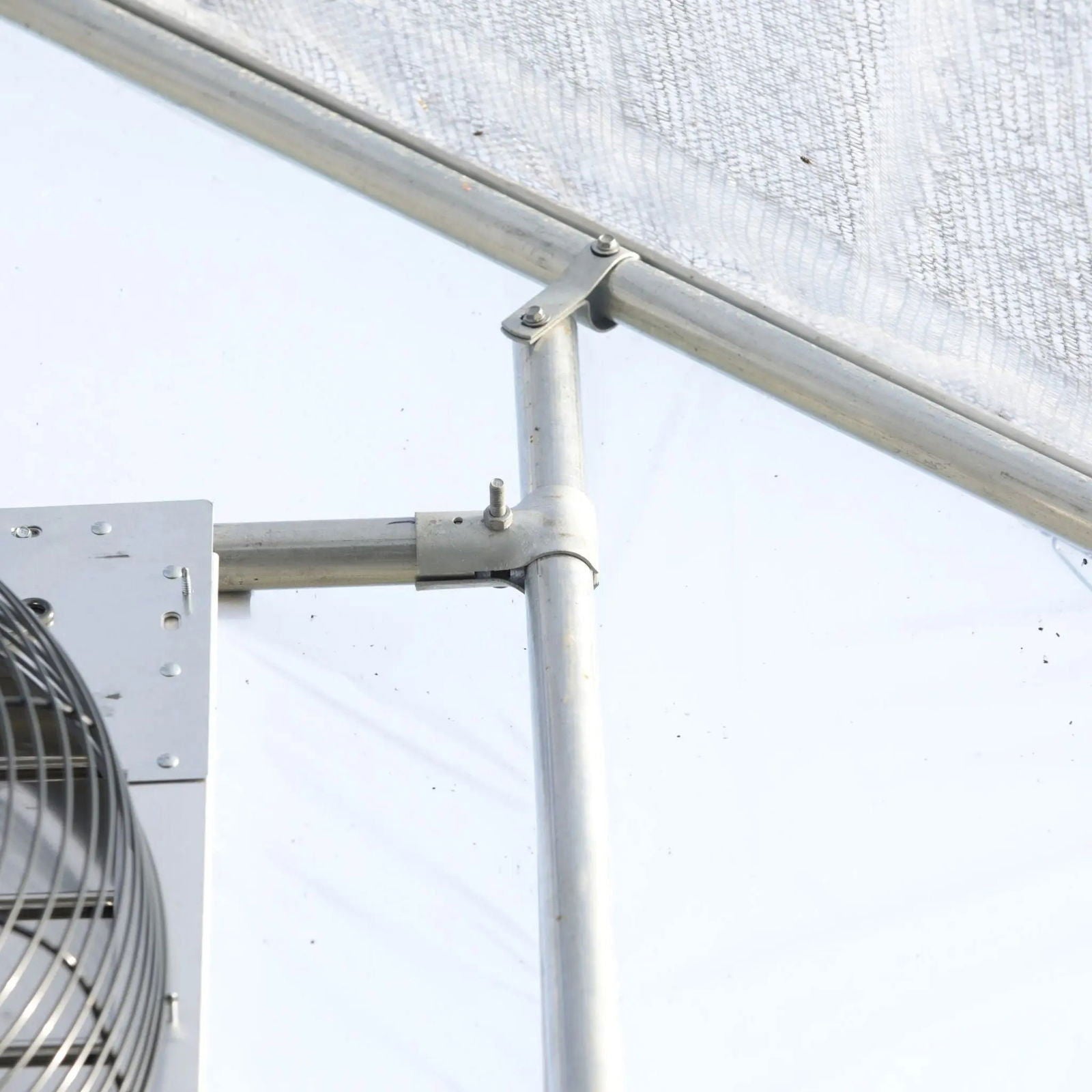 A close-up of Bootstrap Farmer's Greenhouse Shutter Mount Exhaust Fans shows the metal pipe joint and support bracket attached to a hoop house frame, with translucent greenhouse panels in the background.