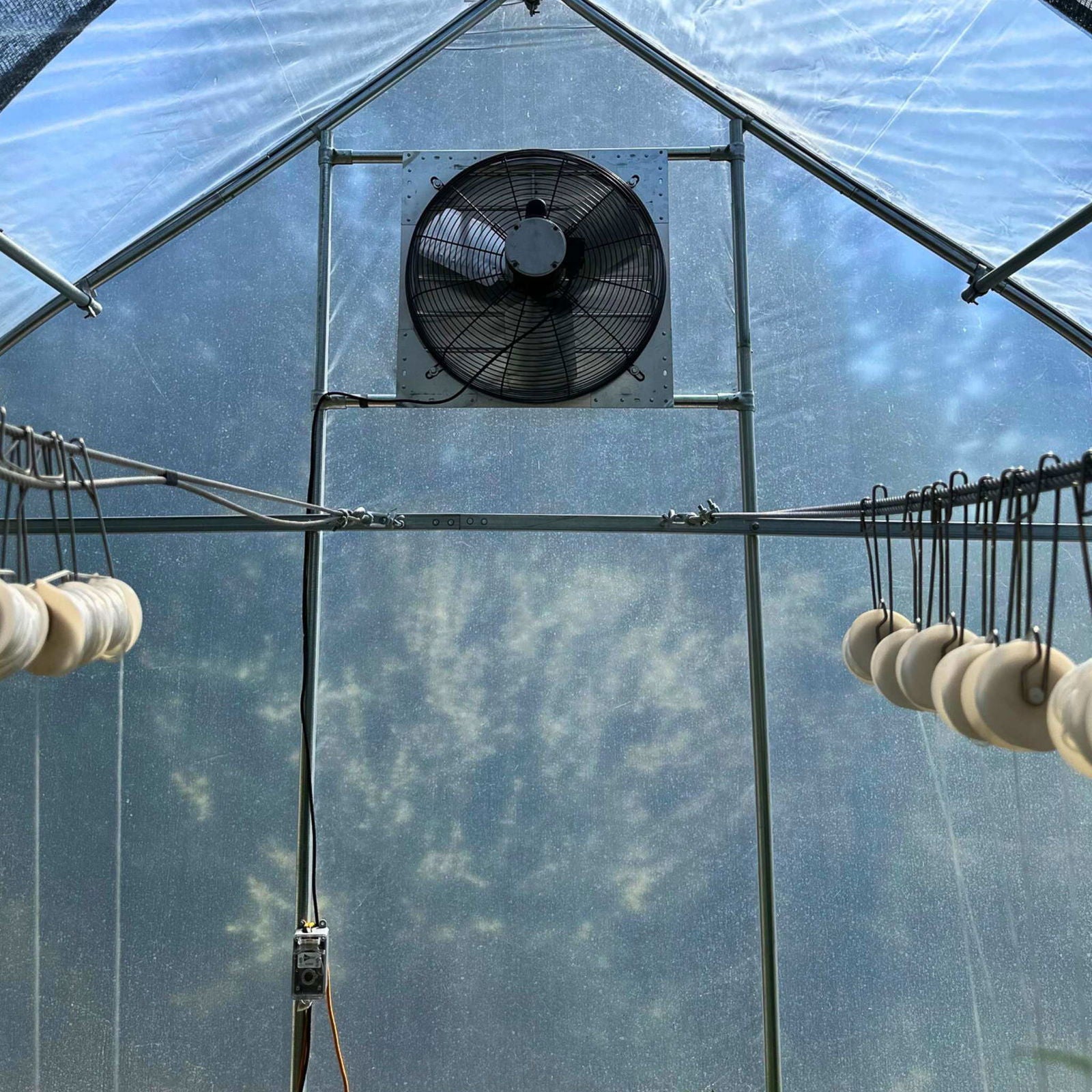A Bootstrap Farmer Greenhouse Shutter Mount Exhaust Fan is installed high on the wall inside a greenhouse, with rows of hanging white items visible as sunlight streams through the translucent walls.
