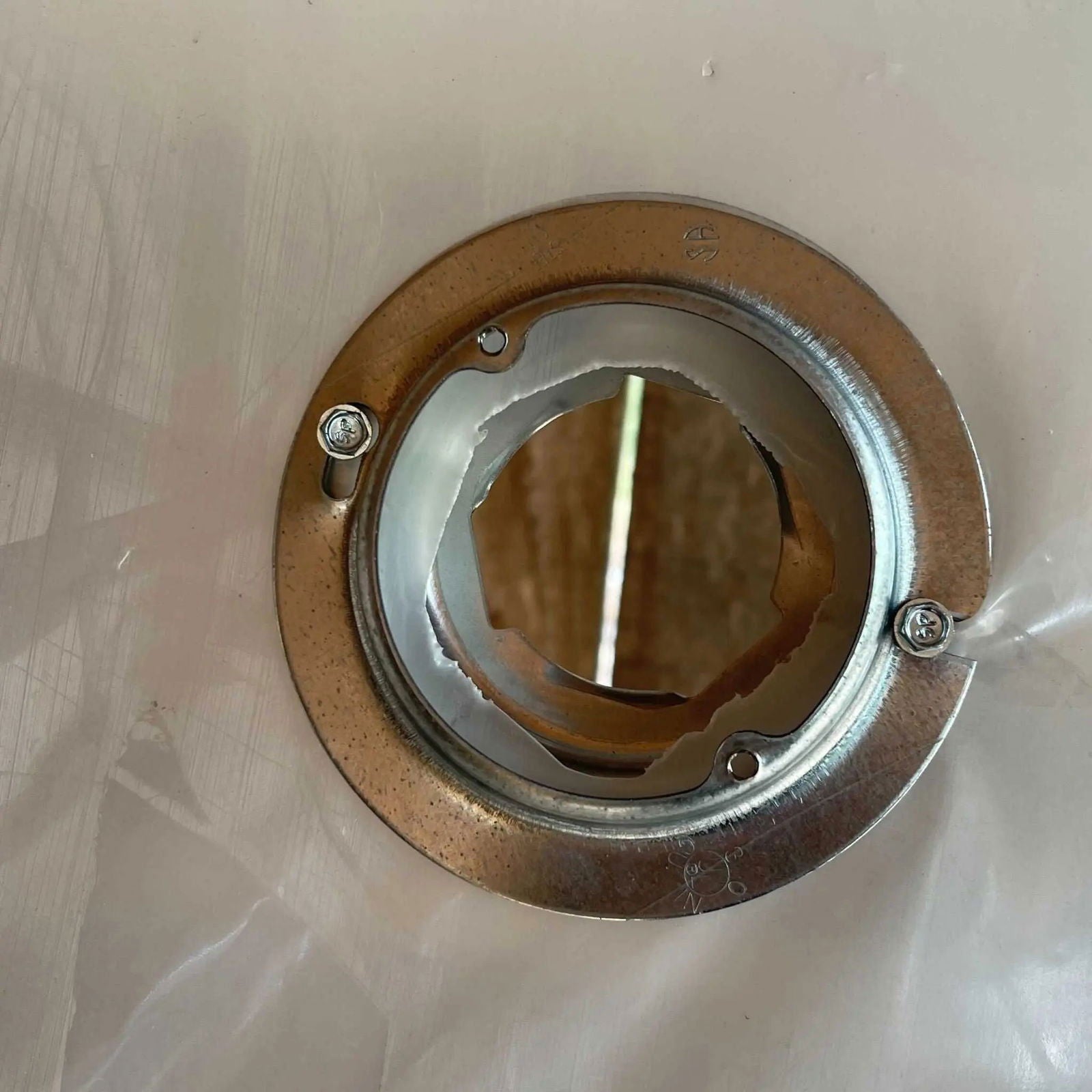 The Bootstrap Farmer Greenhouse Air Inflation Kit features a circular metal flange mounted with three screws, displaying torn insulated greenhouse plastic through the center opening and revealing a wooden surface behind it.