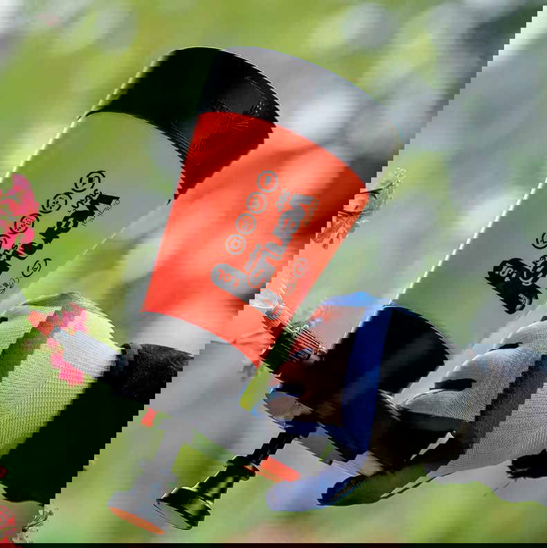 A person holds a red and black reusable coffee cup with a white logo outdoors, while a Bootstrap Farmer Kwazar Venus Handheld Pump Sprayer is visible in the blurred greenery background.