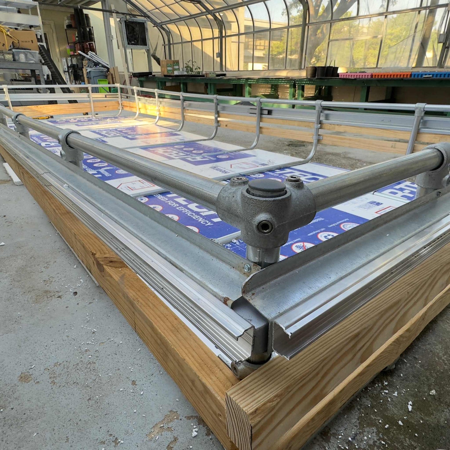 A Bootstrap Farmer Deep Water Culture (DWC) Kit with a wooden raised bed, metal piping framework, and a partially installed plastic cover sits in the greenhouse, indicating a sustainable farming venture. This kit offers a customizable modular system ideal for various gardening projects.