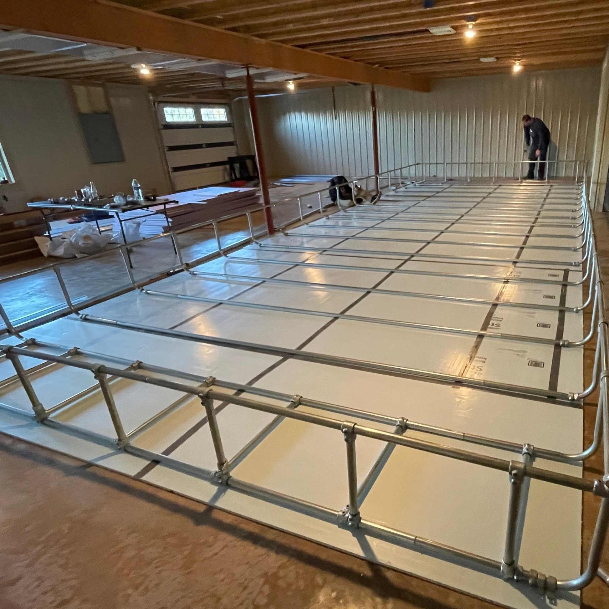 A Bootstrap Farmer DWC Kit is being assembled from metal pipes in a spacious, unfinished basement. A person in the background appears to be constructing the framework for this customizable modular system. Bare walls and floors with materials nearby suggest future sustainable farming projects.