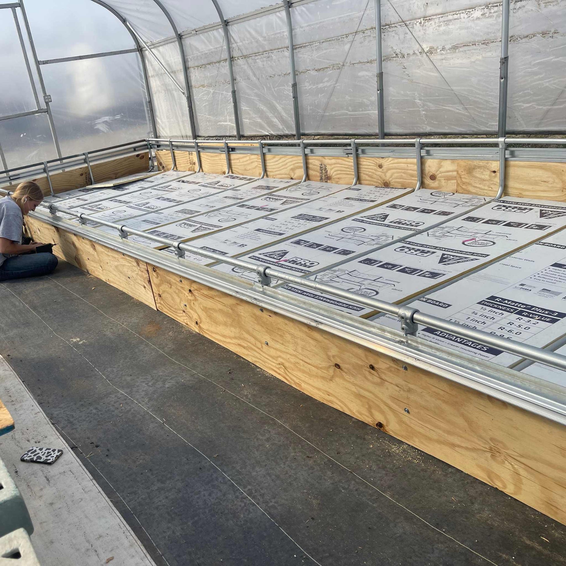 Inside the greenhouse, equipped with a customizable modular system and transparent plastic roof, someone sits by a large rectangular wooden planter box adorned with sustainable farming posters. The setup optimizes light for Bootstrap Farmers Deep Water Culture (DWC) Kit.