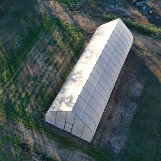 Aerial view of a white fabric greenhouse situated on a grassy field showing tree shadows and a small gated area nearby
