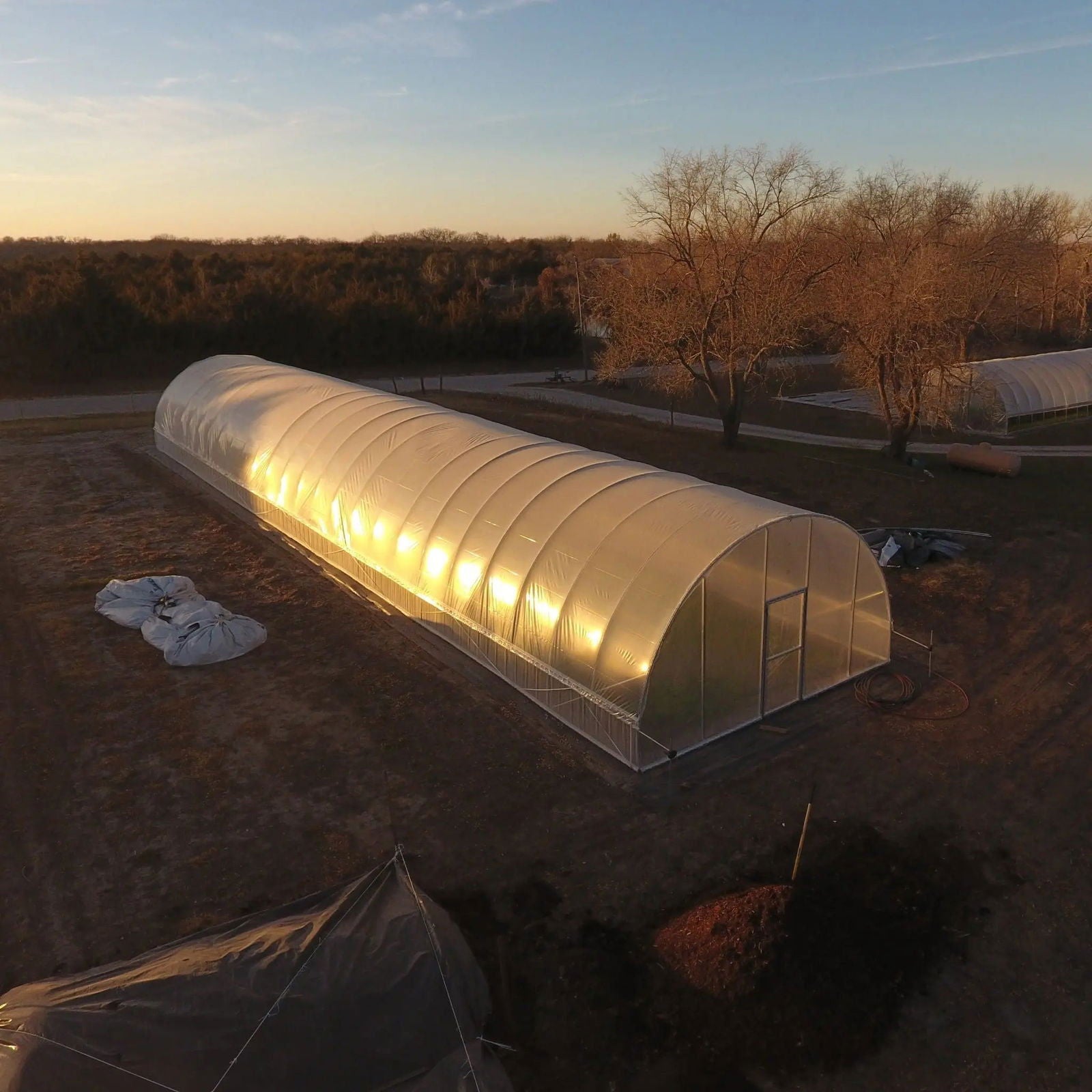Aerial photograph of a large hoop greenhouse on a rural property featuring a golden sunset glow illuminating the translucent plastic, a pile of mulch, and a smaller secondary greenhouse in the background
