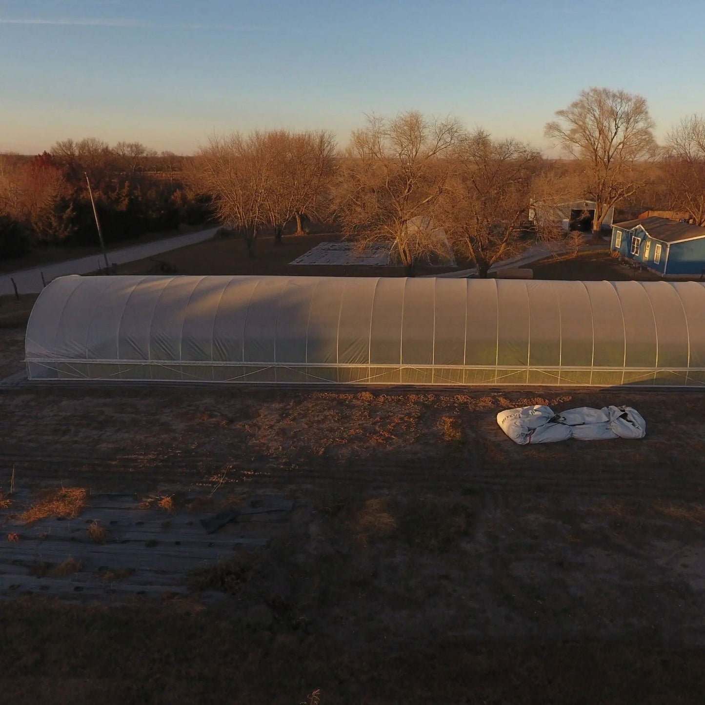 Aerial photograph of a large agricultural greenhouse in a rural landscape featuring a blue house and bare winter trees under a clear sunset sky with a long shadow cast on the greenhouse and several large fabric sacks lying on the ground