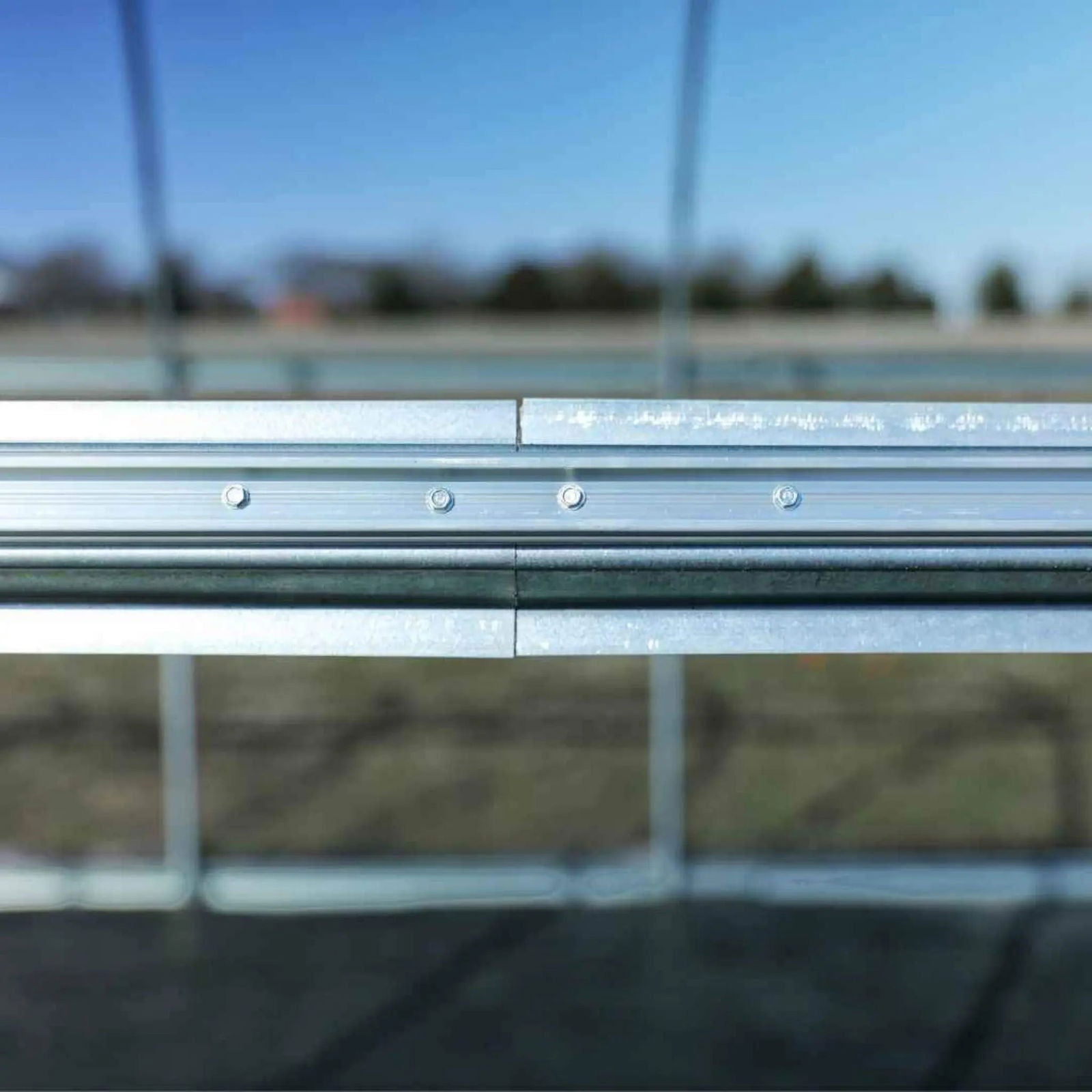 Close-up photograph showing a detail of a metallic greenhouse structure with visible bolts and a blurred background of trees and a clear sky