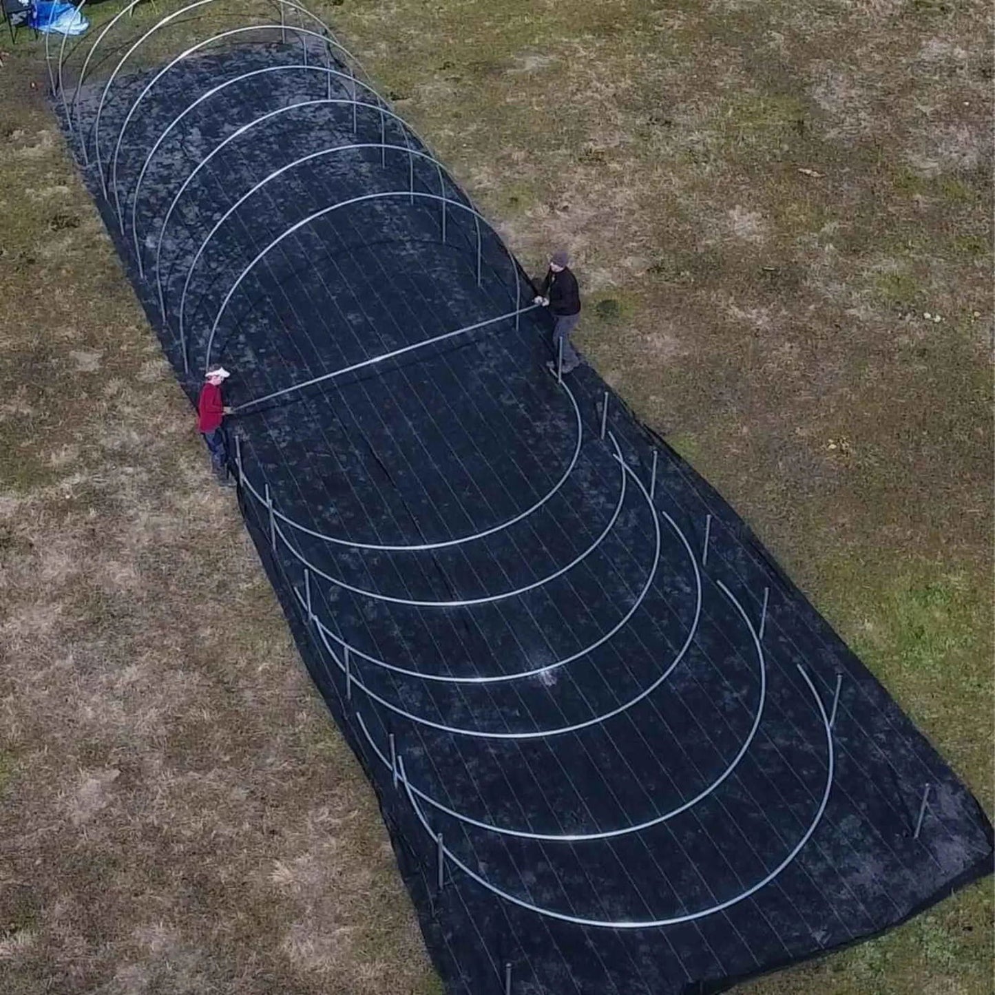 Aerial view of two people constructing a multi-hoop garden structure outdoors on dark landscape fabric using metallic gray hoops and stakes  a red shirt and brown jacket are visible