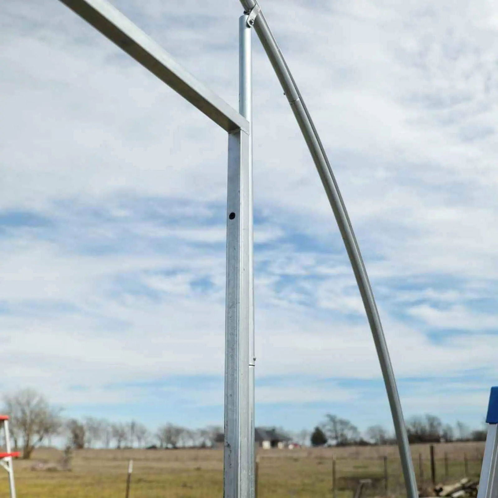 Close-up photo showing galvanized steel framing of a greenhouse under a partly cloudy sky featuring a distant farmstead and bare winter trees with a visible bolt and a slight curve in one of the metal poles
