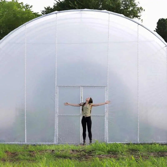 Photograph unknown artist a woman with arms outstretched stands before a large translucent greenhouse in a verdant field featuring a central double door and a curved metallic frame with subtle shadows and lush green grass