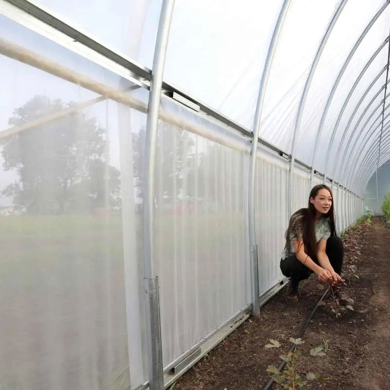 Photograph of a woman tending young plants inside a long, white plastic-covered high tunnel greenhouse with metal framing and visible dark soil showcasing vibrant green growth and a woman with long brown hair