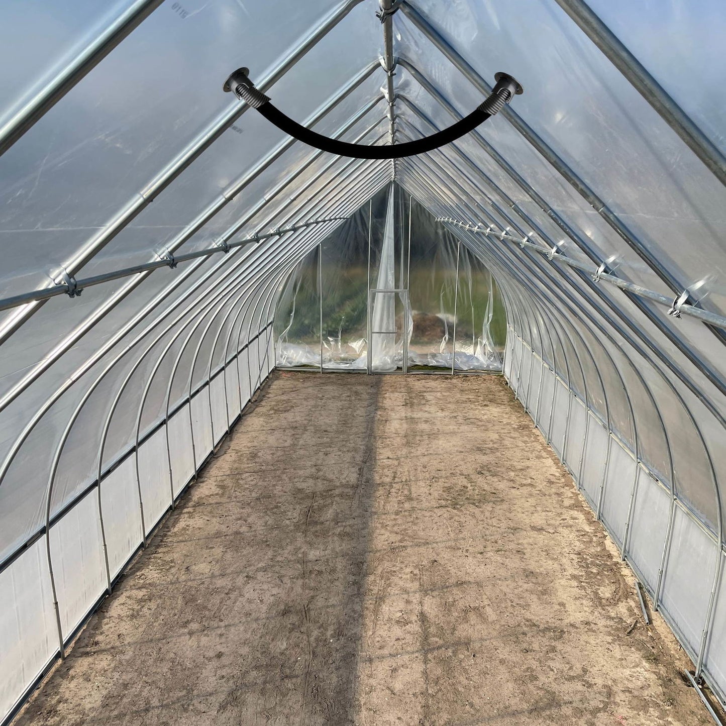 Interior of an empty gothic-style hoop house greenhouse with transparent plastic cover, metal frame, and dirt floor. Sunlight casts shadows, and a Bootstrap Farmer Air Inflation Jumper Hose is attached near the roof.