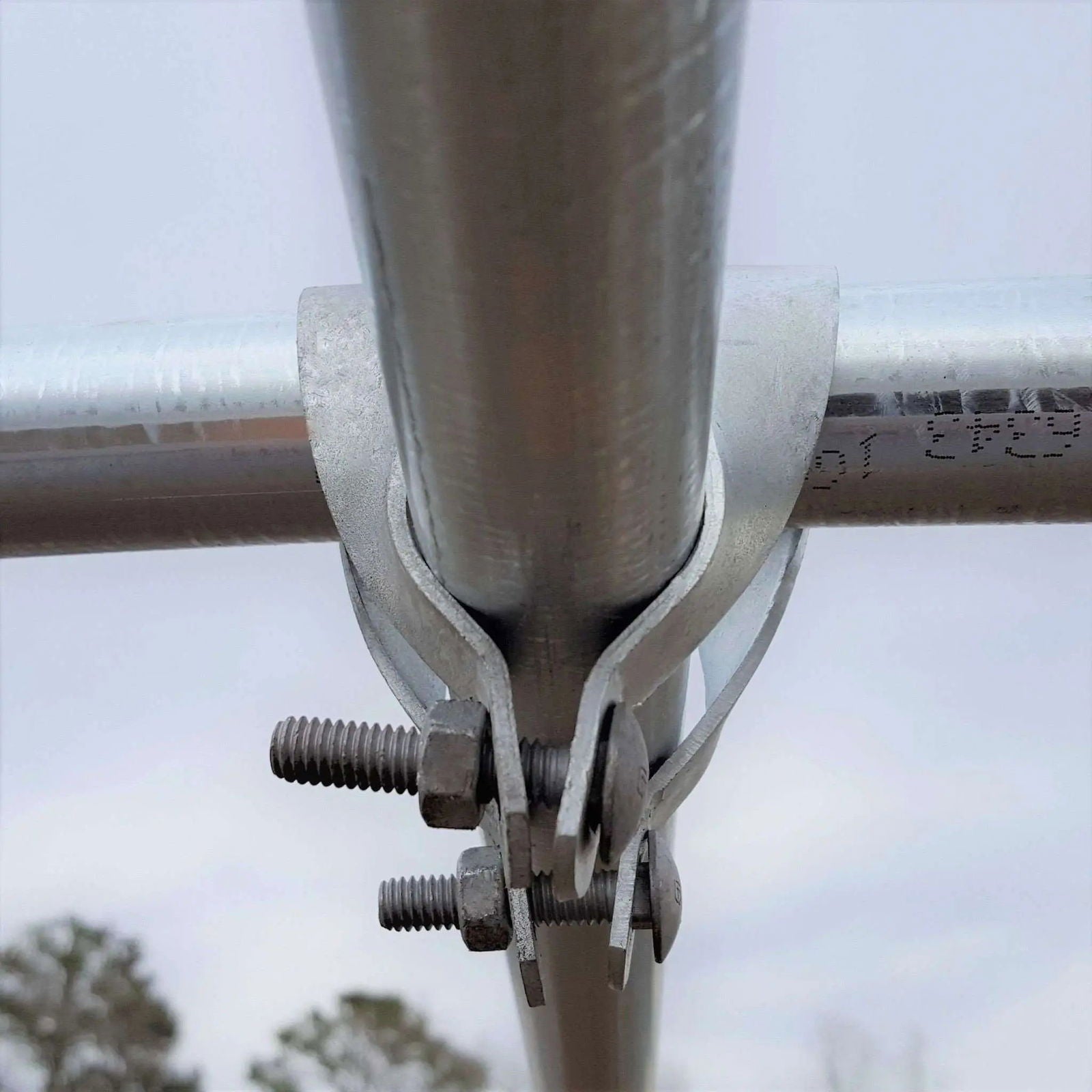 Close-up photograph of a galvanized steel pipe joint with two visible bolts and nuts against a muted sky backdrop showing distant trees