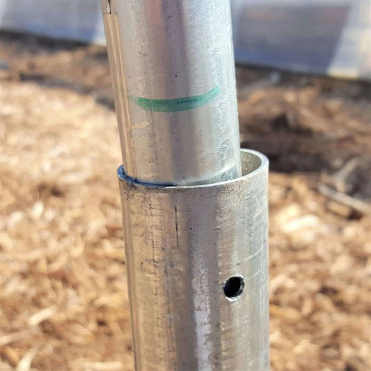 A close-up of Bootstrap Farmer’s Ground Posts - High Tunnel 16 Gauge pipes, one inserted into the other. The outer pipe has a hole, the inner bears a green mark. Brown mulch and a structure are visible in the blurred background.