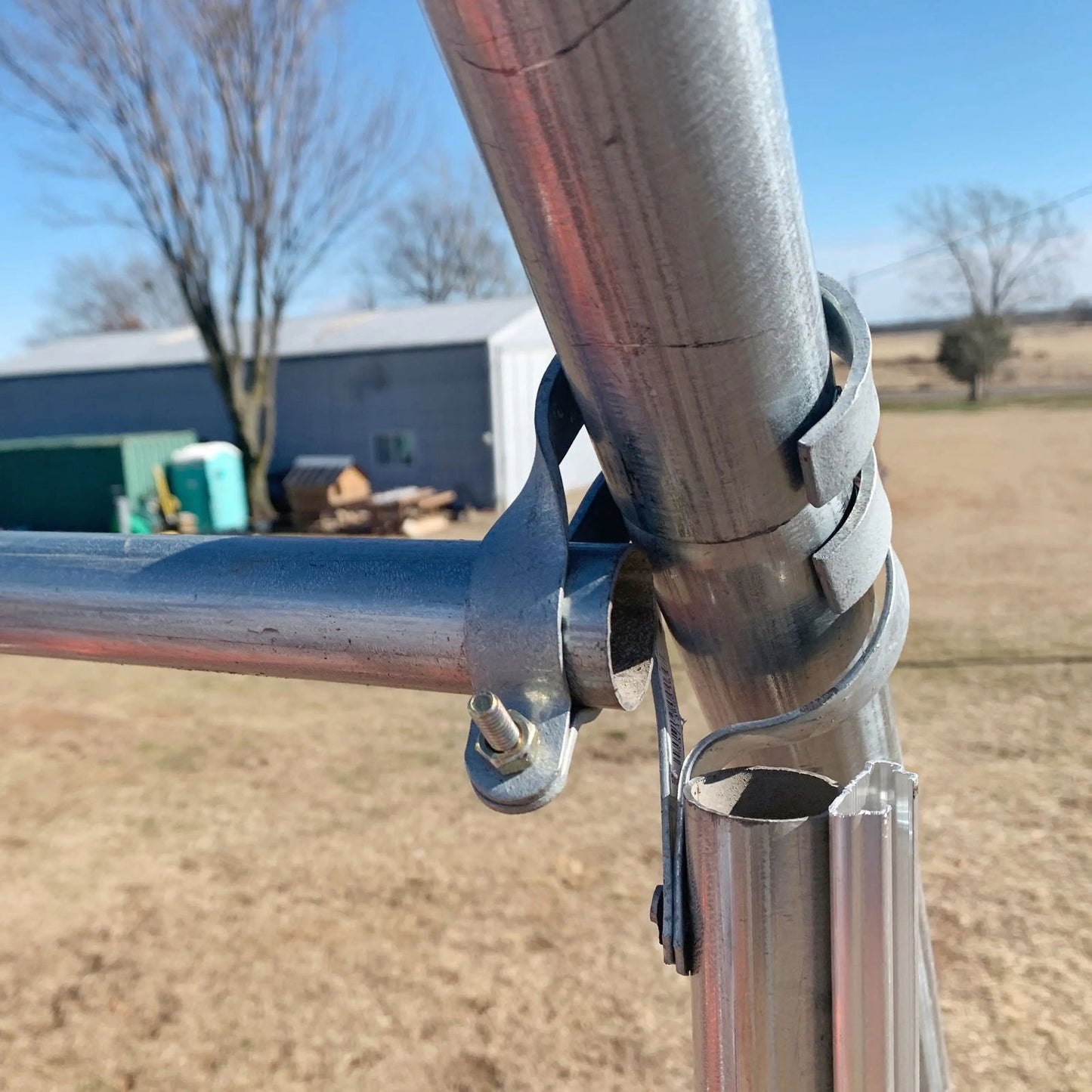 Close-up photograph showing galvanized steel pipe joint construction with a bolt and clamp detail against a rural backdrop featuring a gray barn and leafless trees in winter
