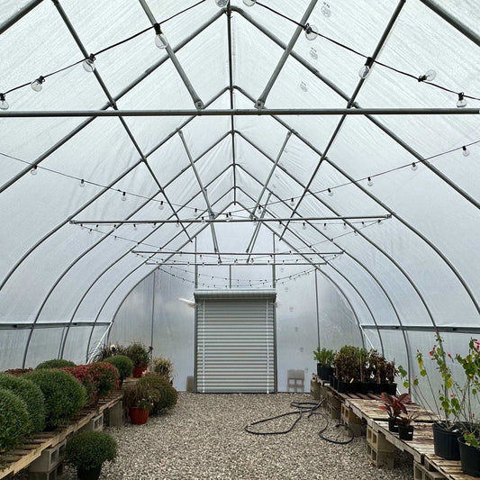 The Truss Kit by Bootstrap Farmer is shown installed in a greenhouse with a gravel floor, potted plants on pallets, and string lights hanging from the arched metal frame. A closed metal door appears at the far end.