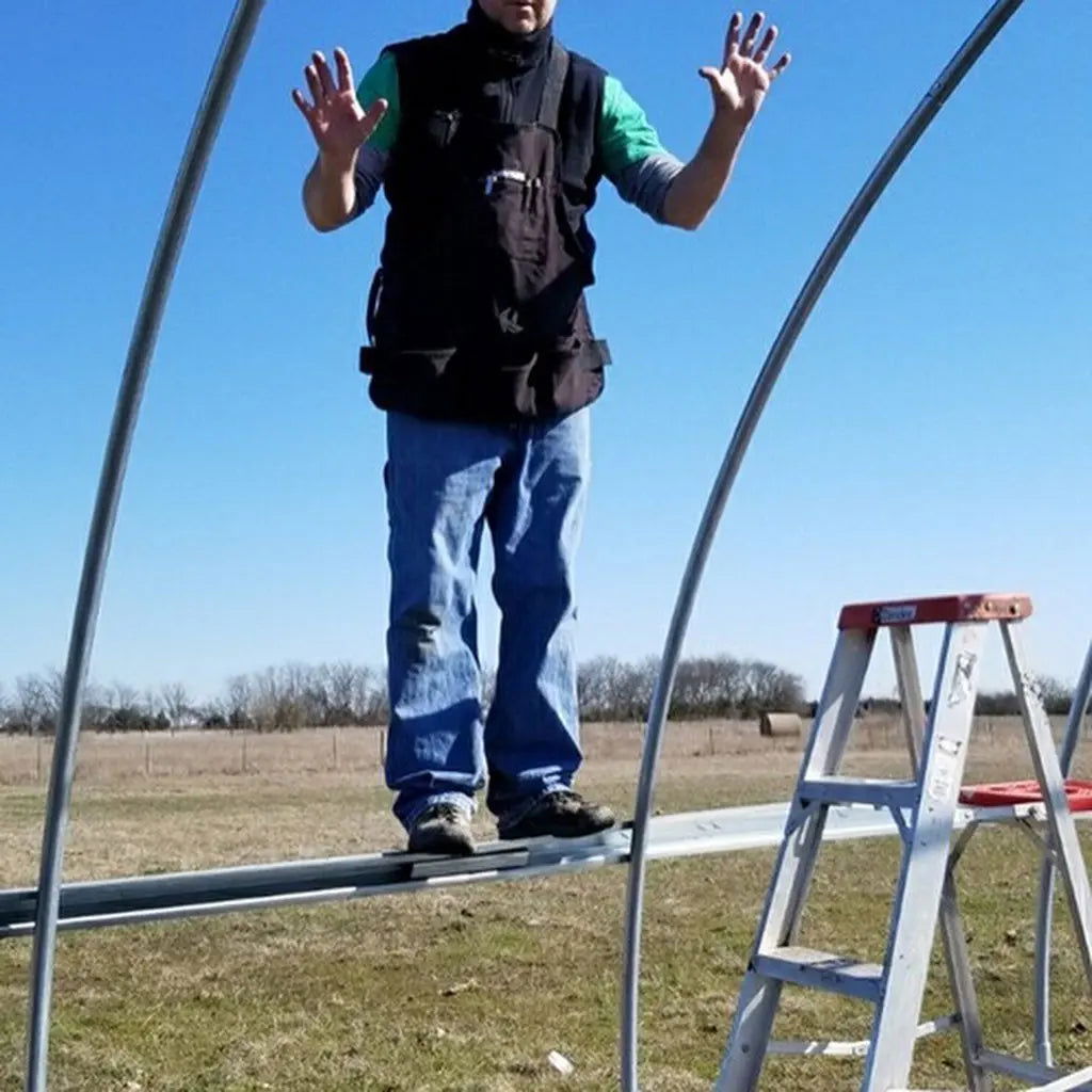 A person stands on a metal beam outdoors beside a step ladder, demonstrating the Bootstrap Farmer Greenhouse Steel Hip and Brace Channel on grass with trees and a clear blue sky in the background.