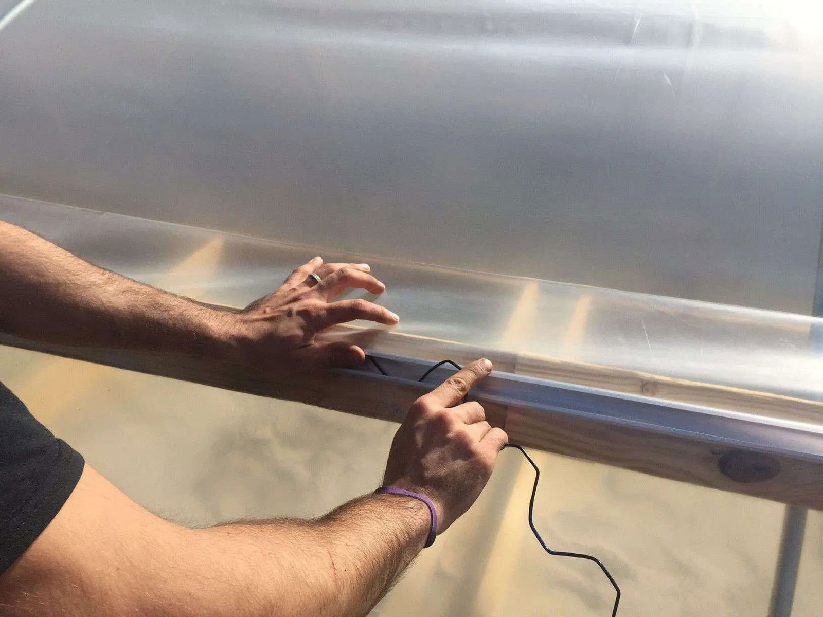 Someone uses Bootstrap Farmer’s Greenhouse PVC Coated Spring Wire & Lock Channel Bundle to secure plastic to a wooden frame, holding the black spring wire against the plastic as sunlight filters through the translucent cover.