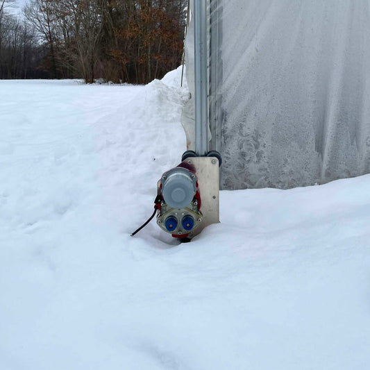 A Bootstrap Farmer Automated Roll-Up Sides Motor is installed at the building's base, partly buried in snow beside a translucent wall—perfect for DIY greenhouse kits or sidewall automation—with snowy trees visible in the background.