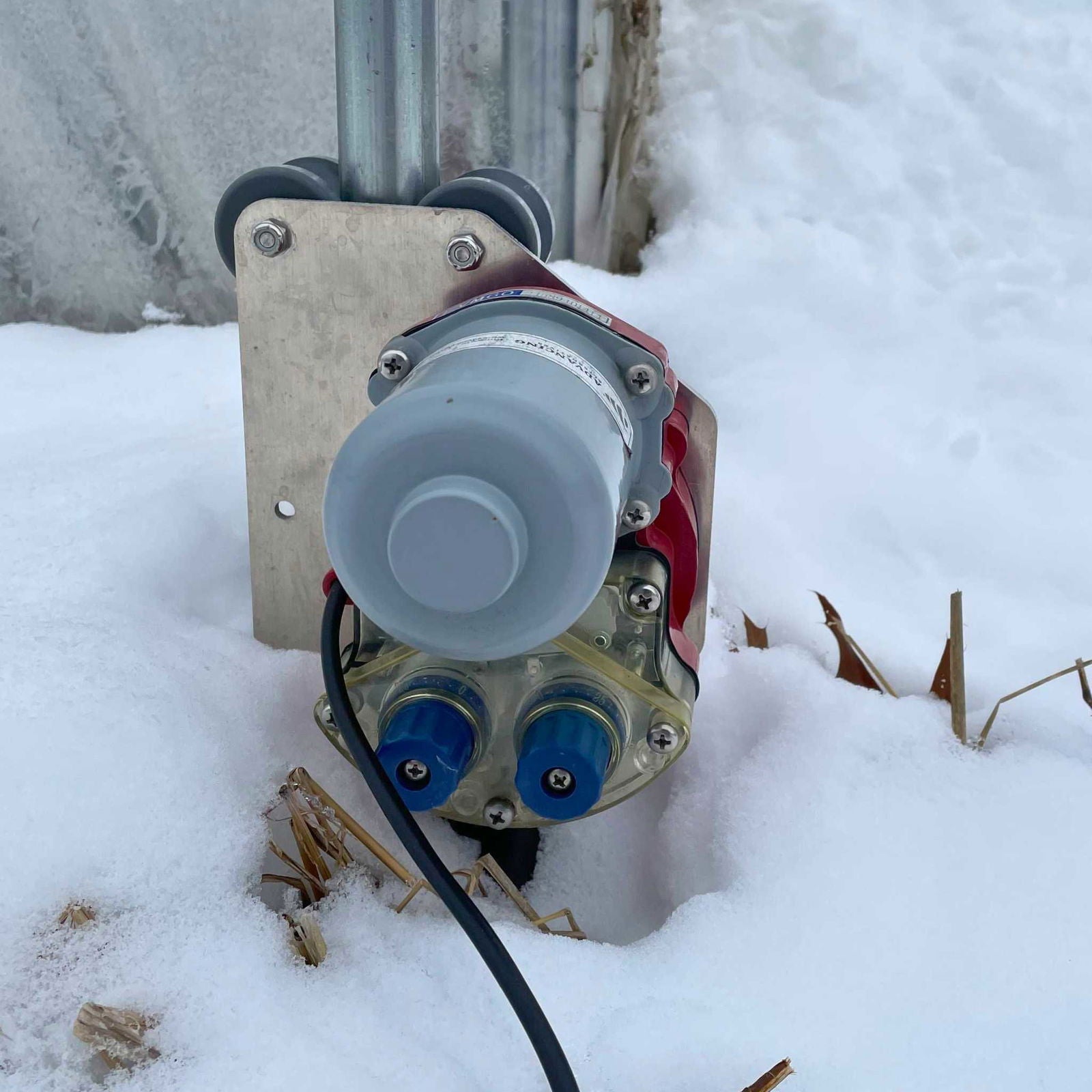 A close-up of the Bootstrap Farmer Automated Roll-Up Sides Motor with wires, mounted on a metal bracket and partially covered by snow—perfect for DIY greenhouse kits or sidewall automation solutions.