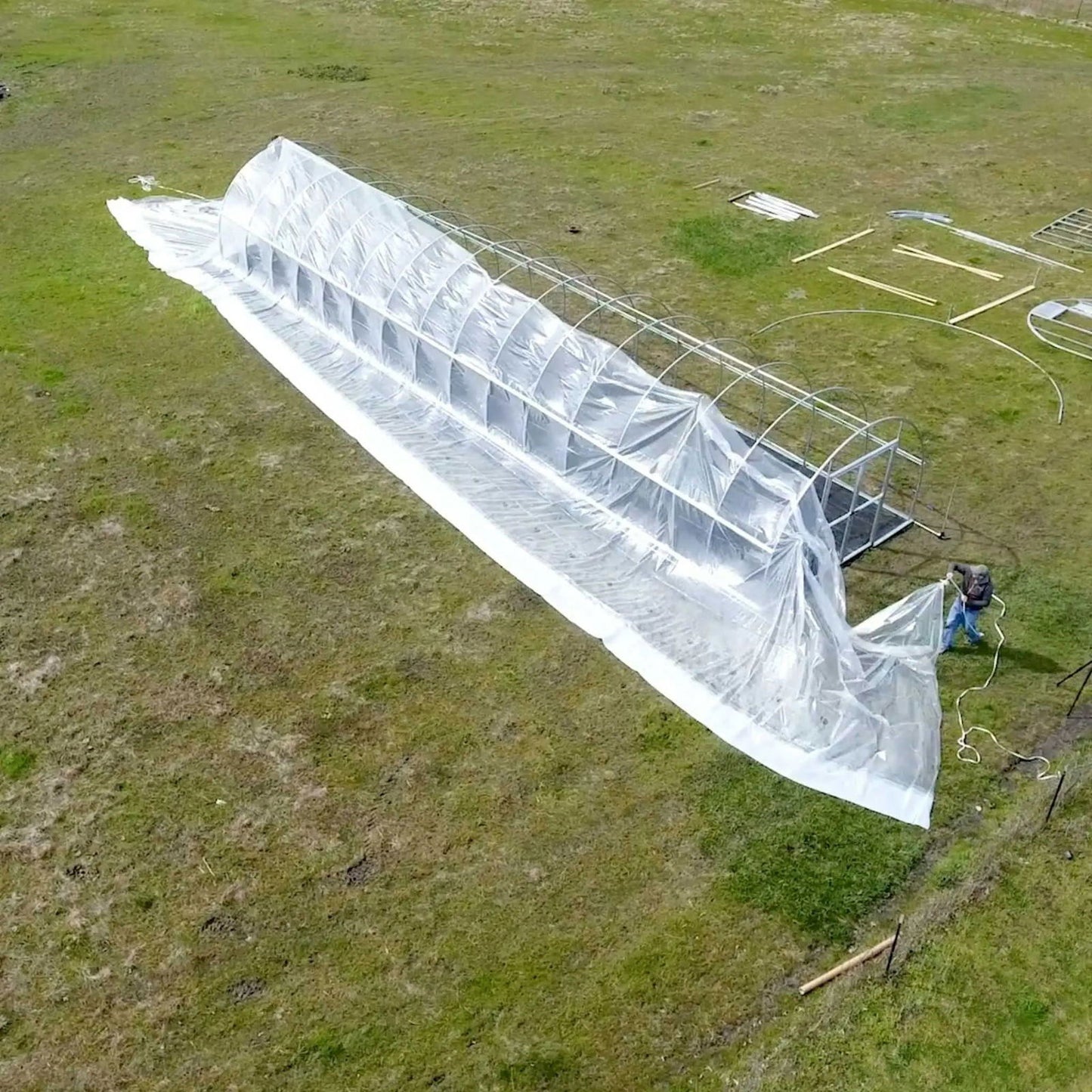 Aerial view of a person assembling a long, translucent plastic greenhouse in a grassy field featuring metal framing, a partially draped plastic cover, and scattered construction materials