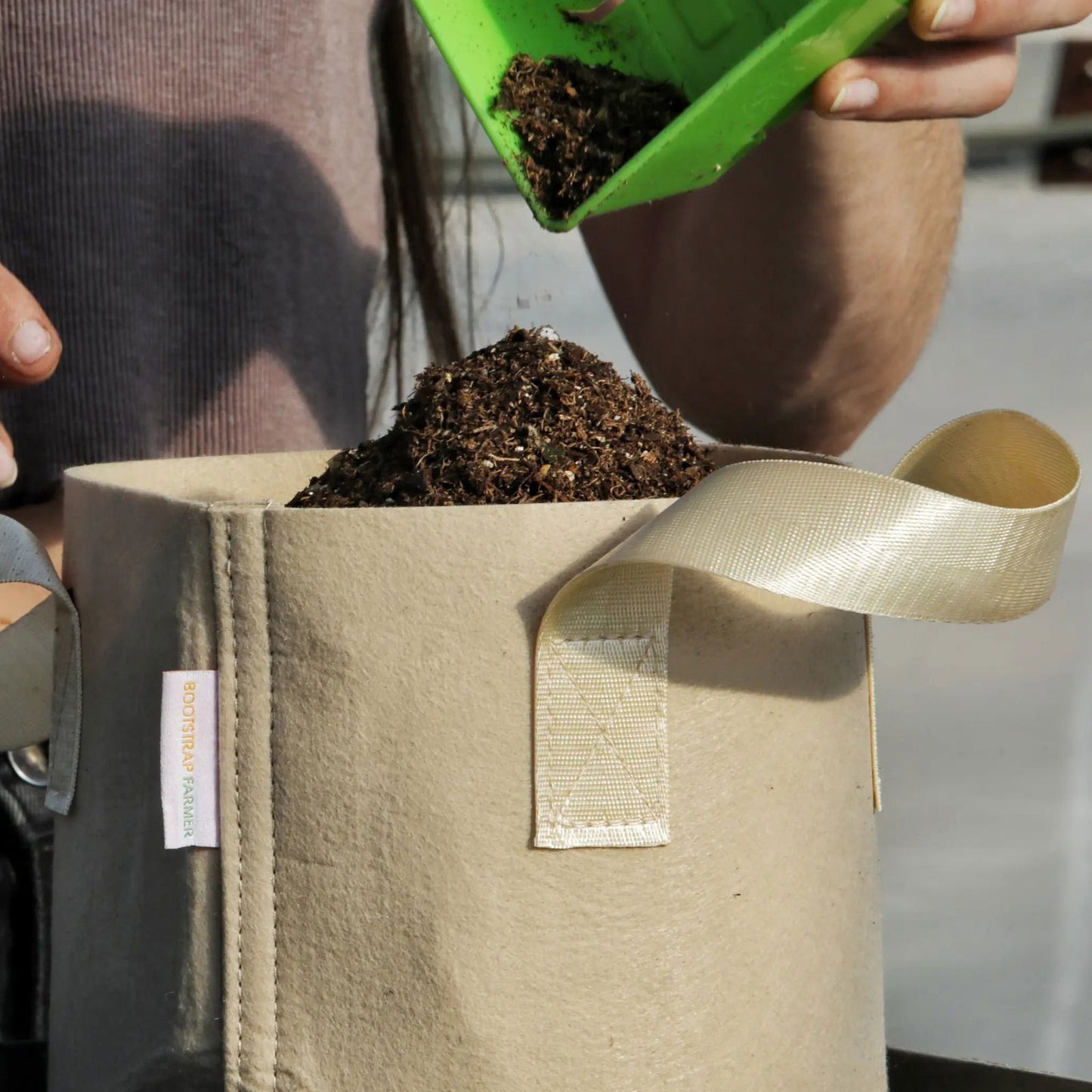 Bootstrap Farmer photograph showing a person filling a beige fabric grow bag with dark brown soil using a bright green scoop the bag features a beige fabric handle and the Bootstrap Farmer logo is visible
