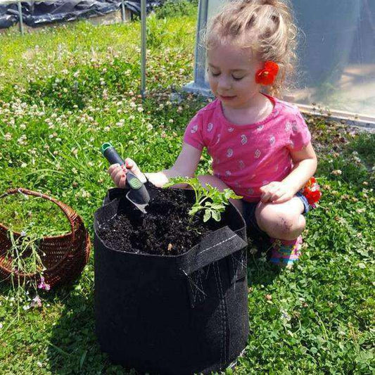 Photograph of a young girl planting a tomato seedling in a black fabric grow bag using a small trowel in a garden with clover and a wicker basket colorful flowers and a greenhouse in the background
