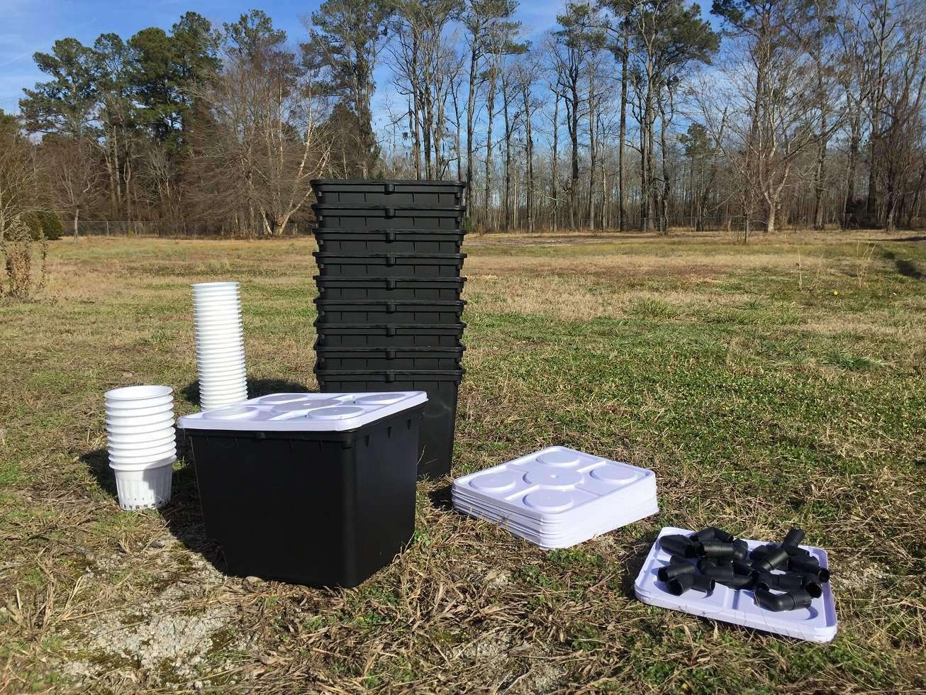 Photograph of hydroponic gardening equipment in a rural field featuring stacked black containers white net pots and black connectors against a backdrop of tall trees
