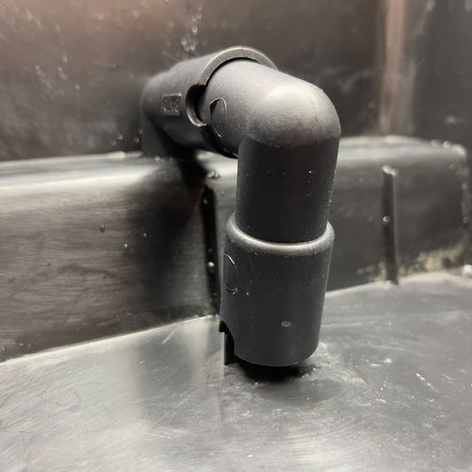 Close-up photo of a matte black plastic plumbing fitting with a 90-degree bend and visible markings  attached to a dark gray surface showing subtle scratches and water droplets
