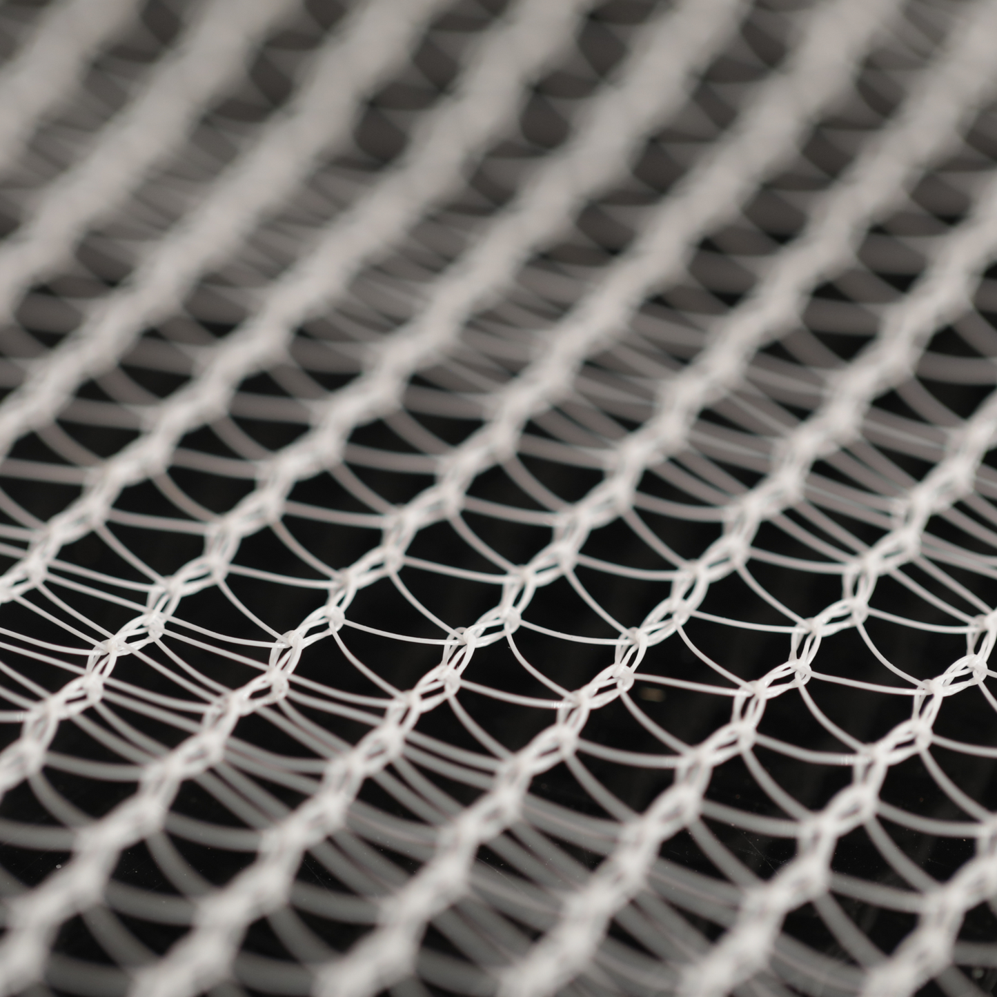 Close-up photograph of a white plastic netting with a diamond pattern showing intricate knots and subtle shadows against a dark background
