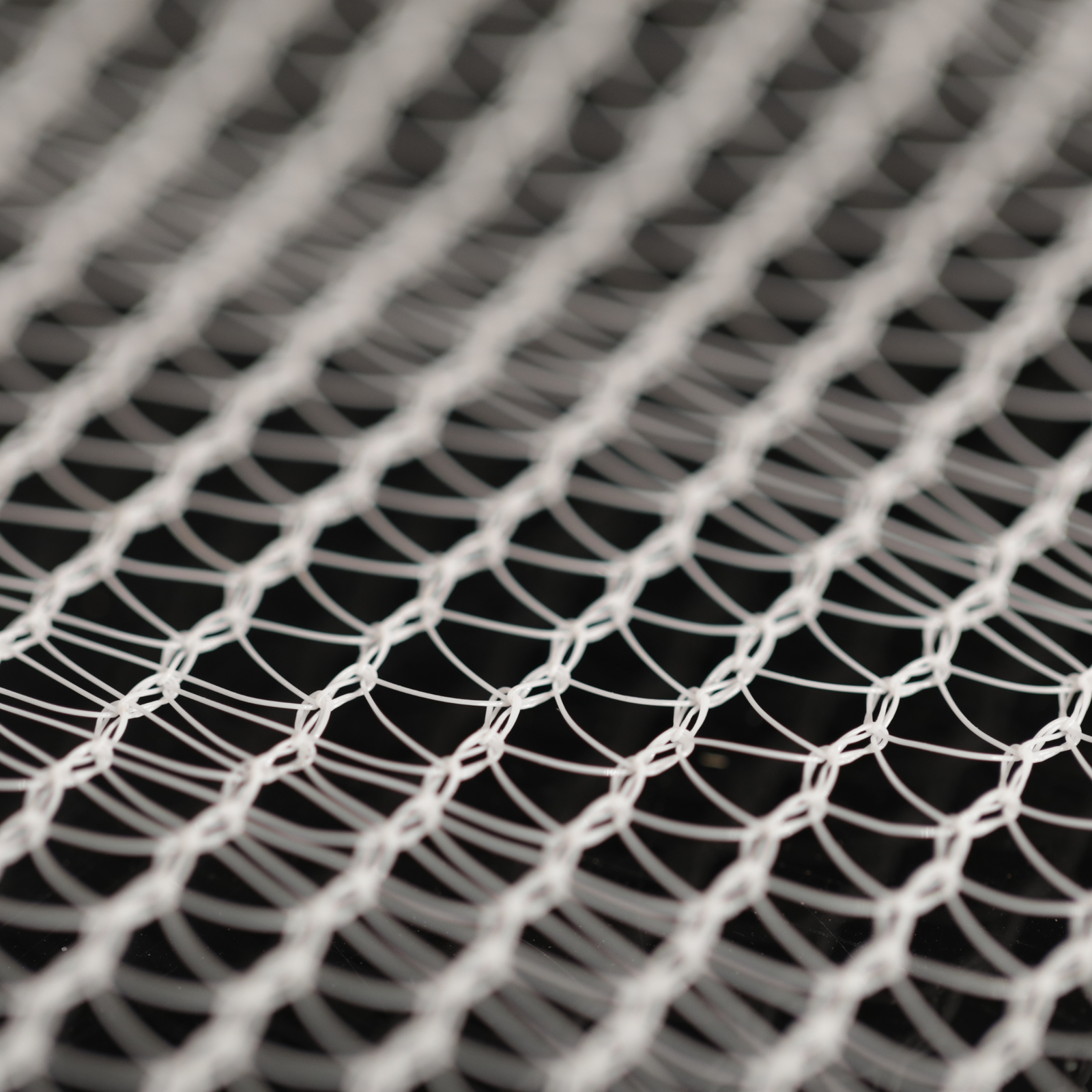 Close-up photograph of a white plastic netting with a diamond pattern showing intricate knots and subtle shadows against a dark background
