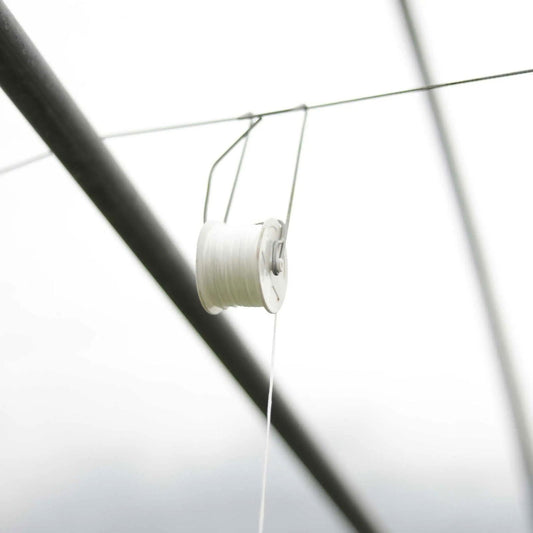 A spool of white thread unwinds downward from a Bootstrap Farmer Tomato Roller Hook, hanging by metal hooks as part of a greenhouse trellis system against a bright background.