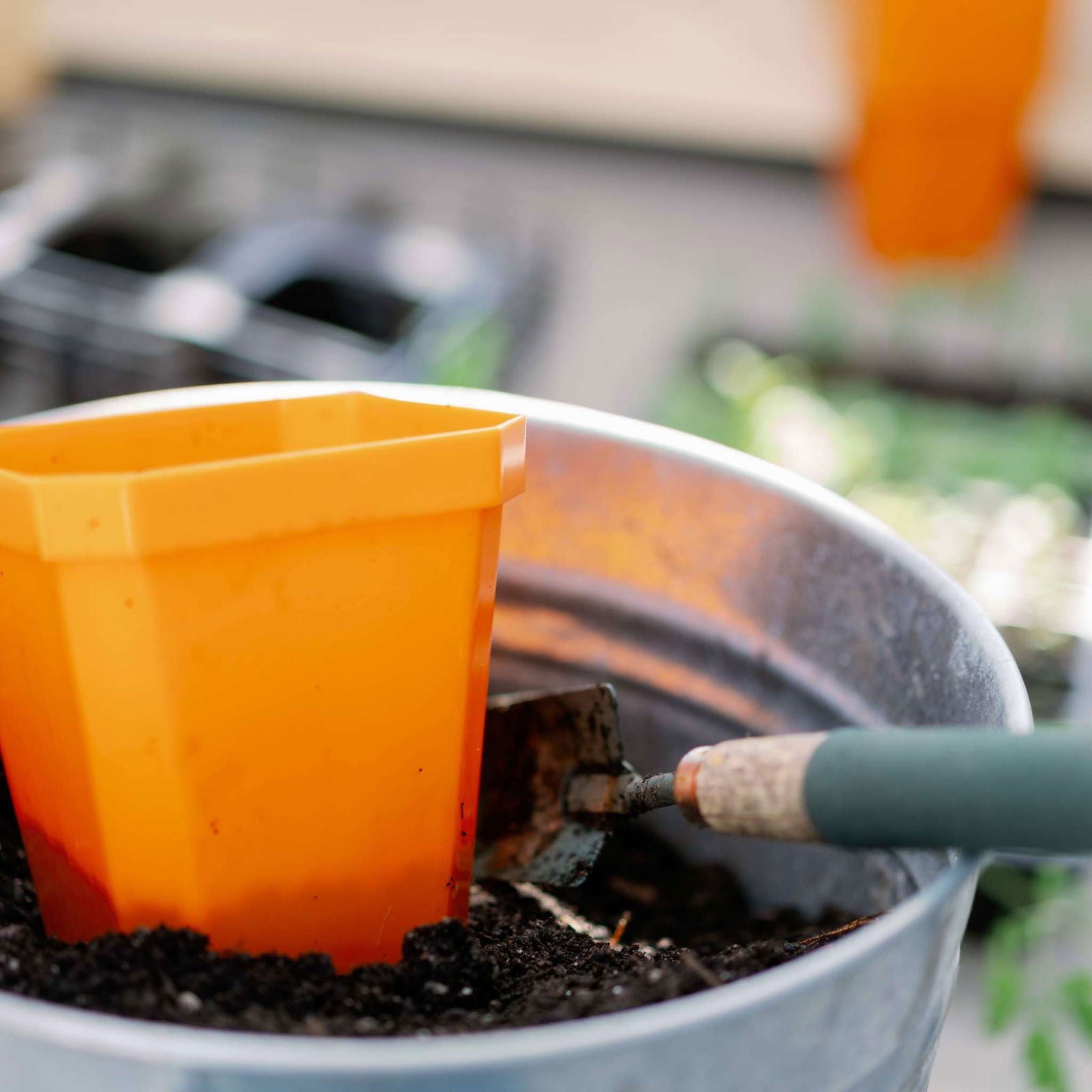 A 5" Heavy Duty Seed Starting Pot by Bootstrap Farmer sits in a metal container with soil, a gardening trowel nearby. Blurred gardening supplies and green seedlings appear in the background.