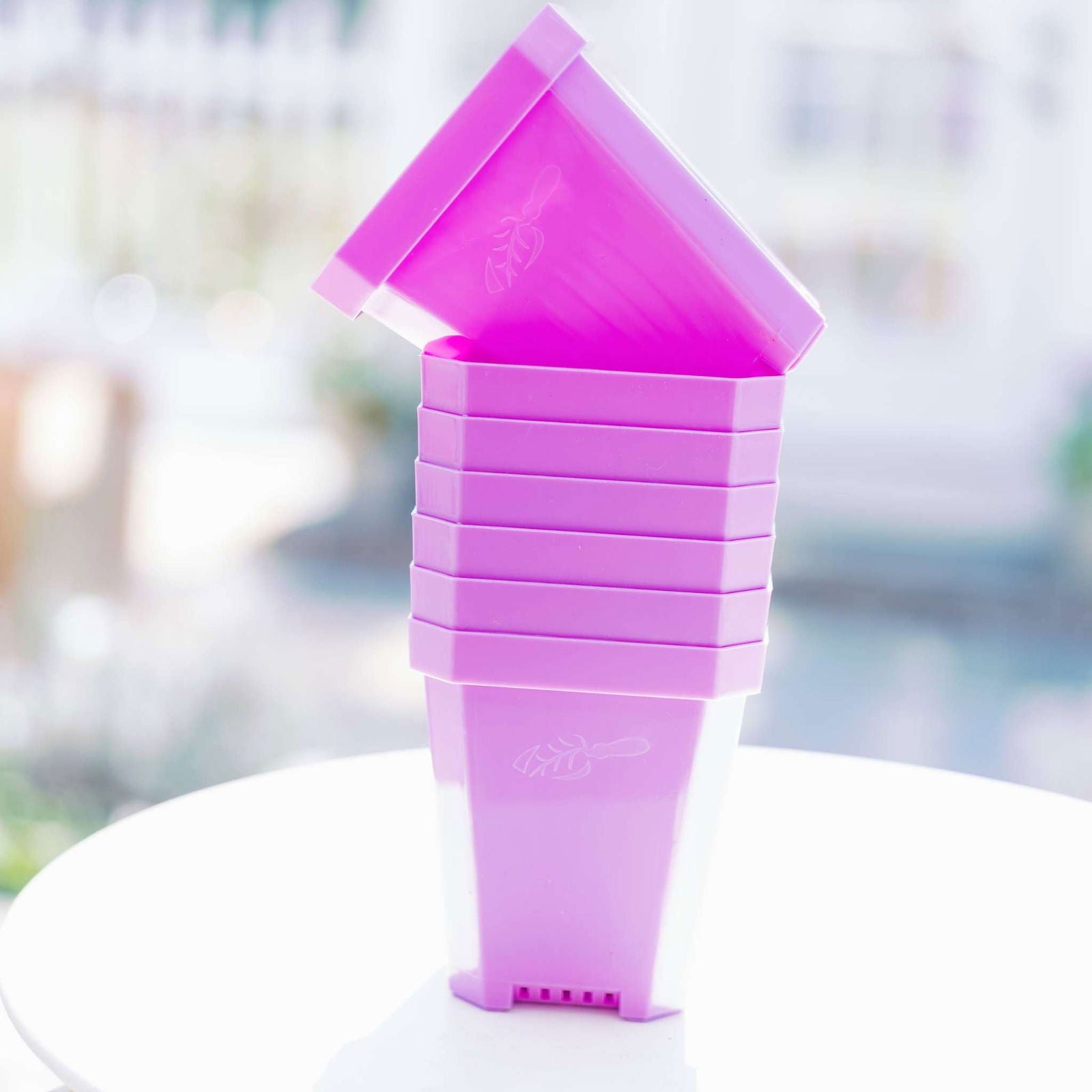 A stack of six Bootstrap Farmer 5" Heavy Duty Seed Starting Pots in bright pink sits on a white surface. The top pot is tilted, showing the starter pots’ subtle embossed leaf design.