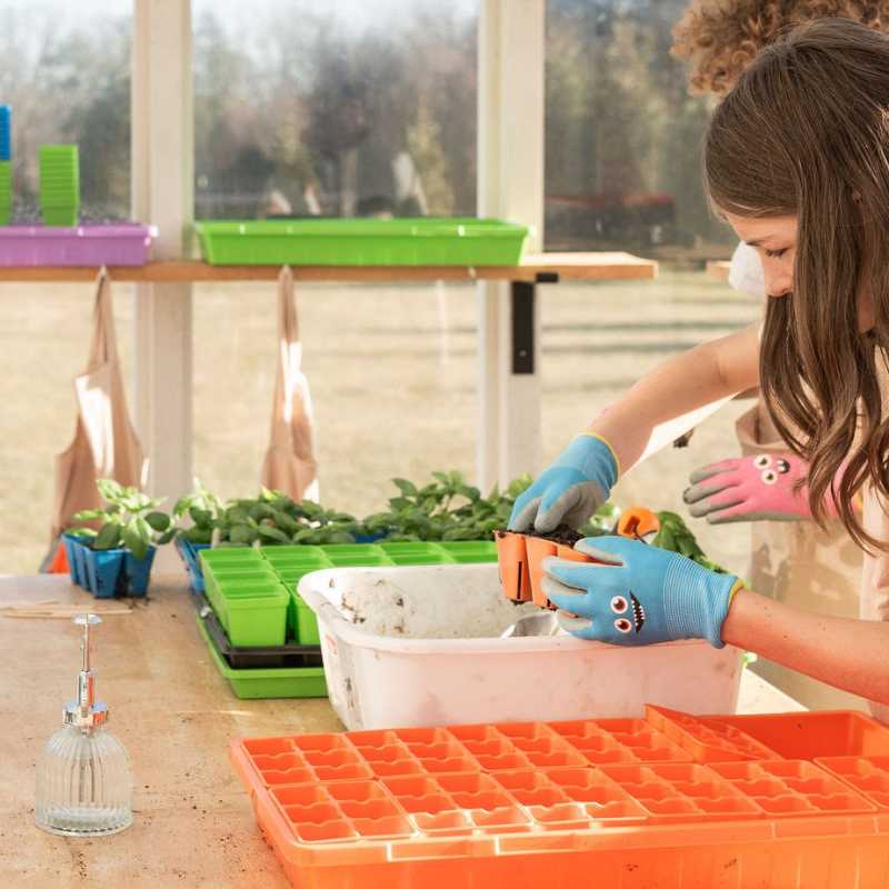 Photograph young girl in greenhouse planting seedlings in orange and bright blue gloves using orange seed starter trays near colorful seed trays and a white plastic container