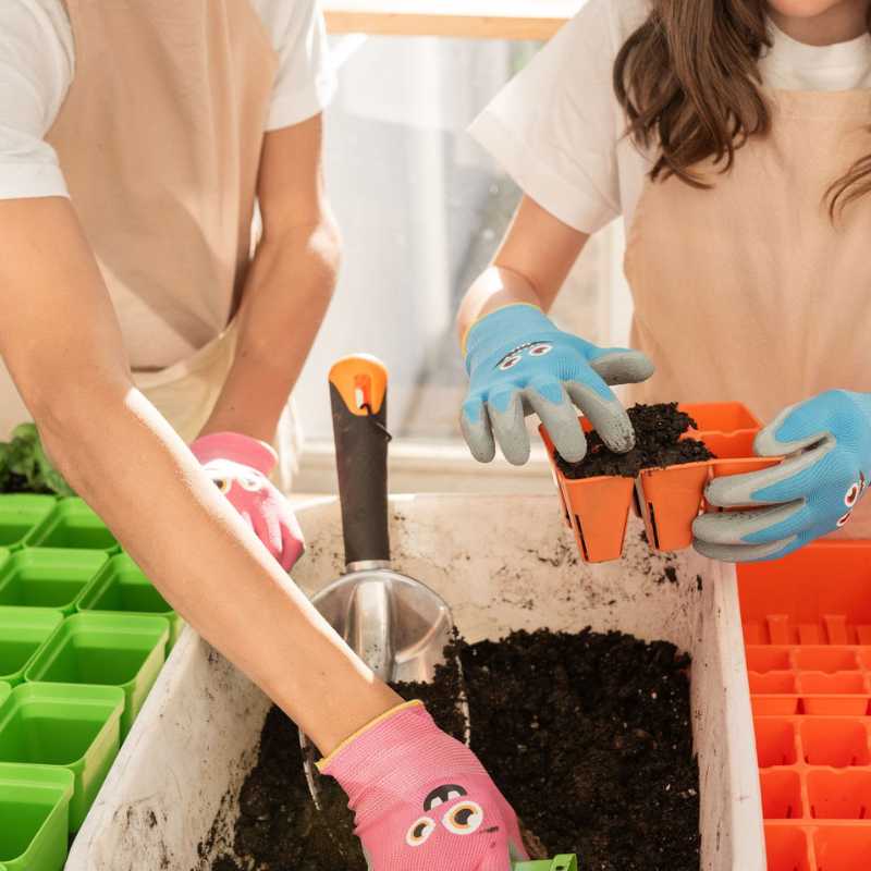 Photographic print of two people planting seedlings in orange seed starter trays using colorful gardening gloves and a metal scoop in a white container filled with dark soil