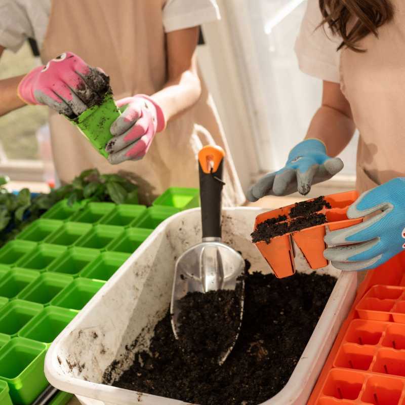 Photograph of two people gardening in a greenhouse using colorful seed starter trays and gloves planting soil into orange and green containers amongst bright green seedling trays and a metal scoop