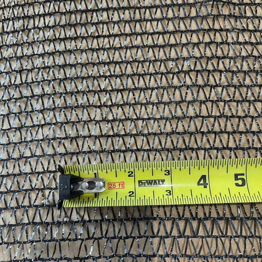 Close-up photograph of black shade cloth with a DeWalt tape measure showing a portion of the material, featuring a repeating triangular pattern, subtle silver highlights, and a textured surface