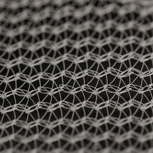 Close-up photograph of a detailed white knotted net-like texture with an intricate pattern of loops and lines against a dark background showing depth and shadows