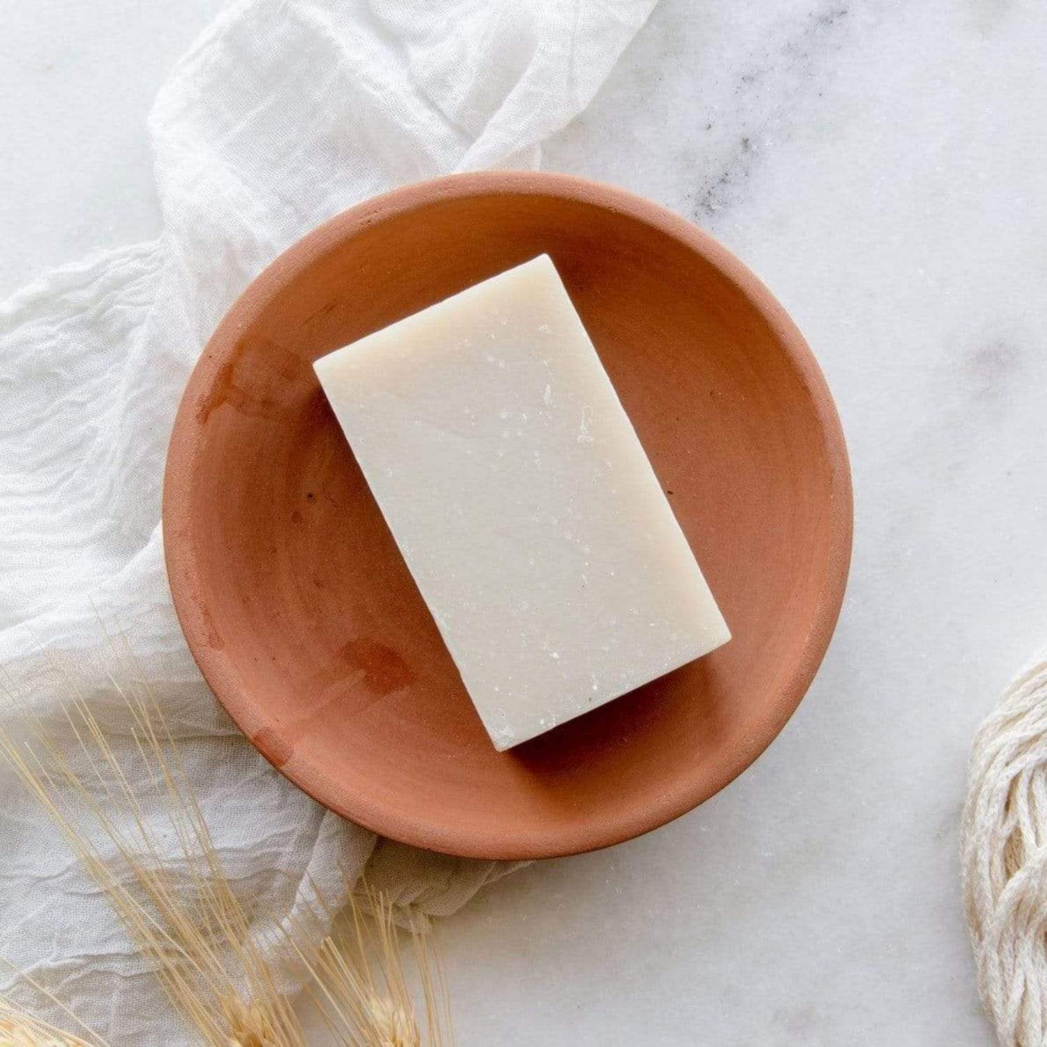 A rectangular 4oz all natural, vegan Soap Bar from ZeroWasteStore.com rests in a terracotta dish on marble, with a white cloth and wheat stems nearby. Available in 6 scents, this plastic-free soap is perfect for face and body.