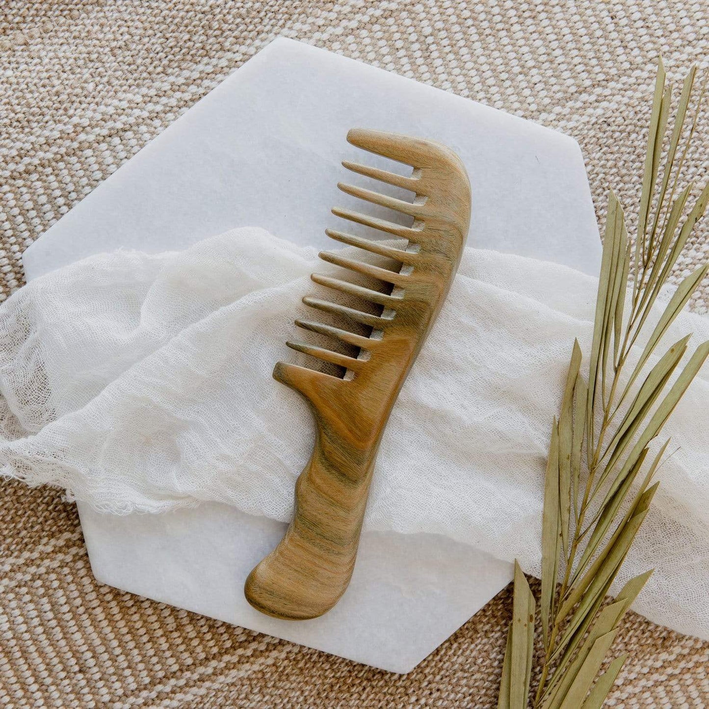 The Sandalwood Wide Tooth Comb by ZeroWasteStore.com, made from 100% wood and plastic free, sits on a white cloth atop a hexagonal marble tray with dried leaves nearby, creating a touch of zero waste elegance.