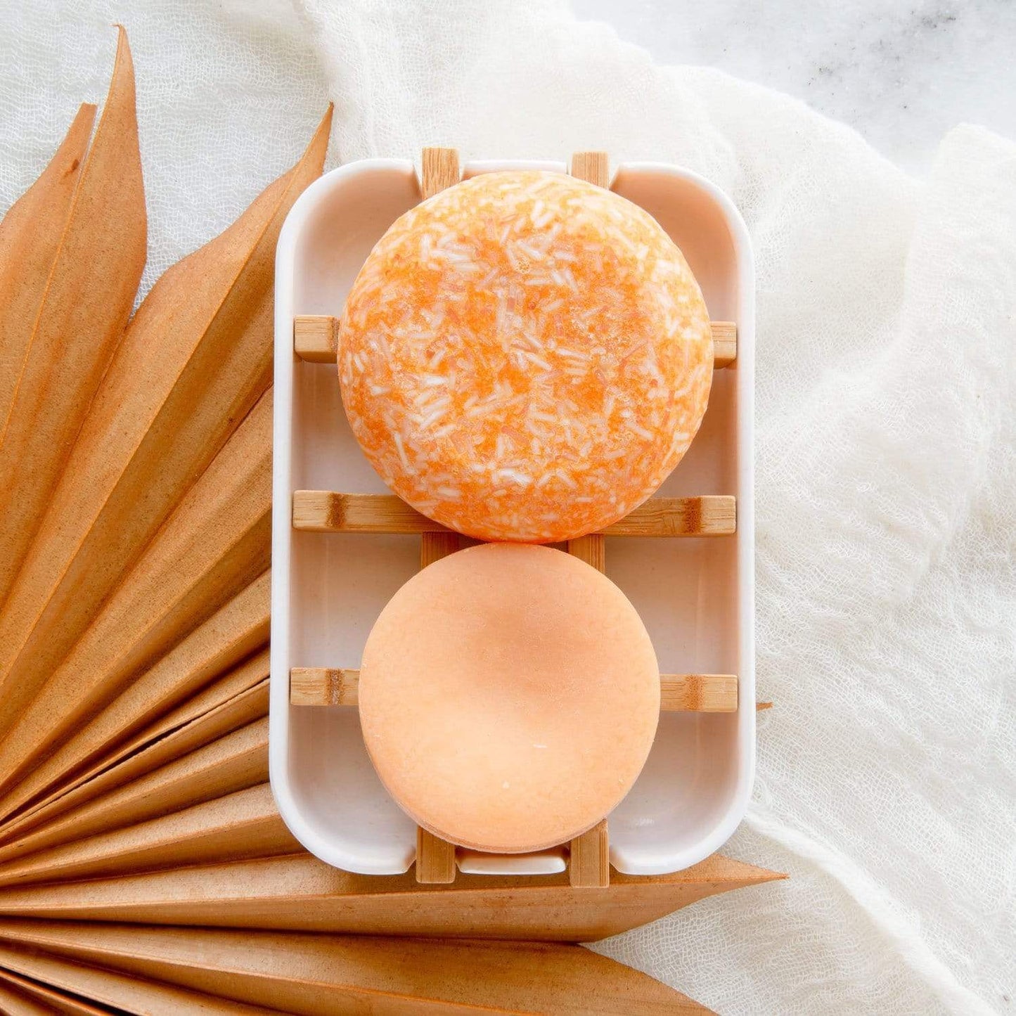 Two round orange soap bars rest on the Bamboo Soap Dish from ZeroWasteStore.com, set in a white tray with a dried palm leaf and white fabric in the background.