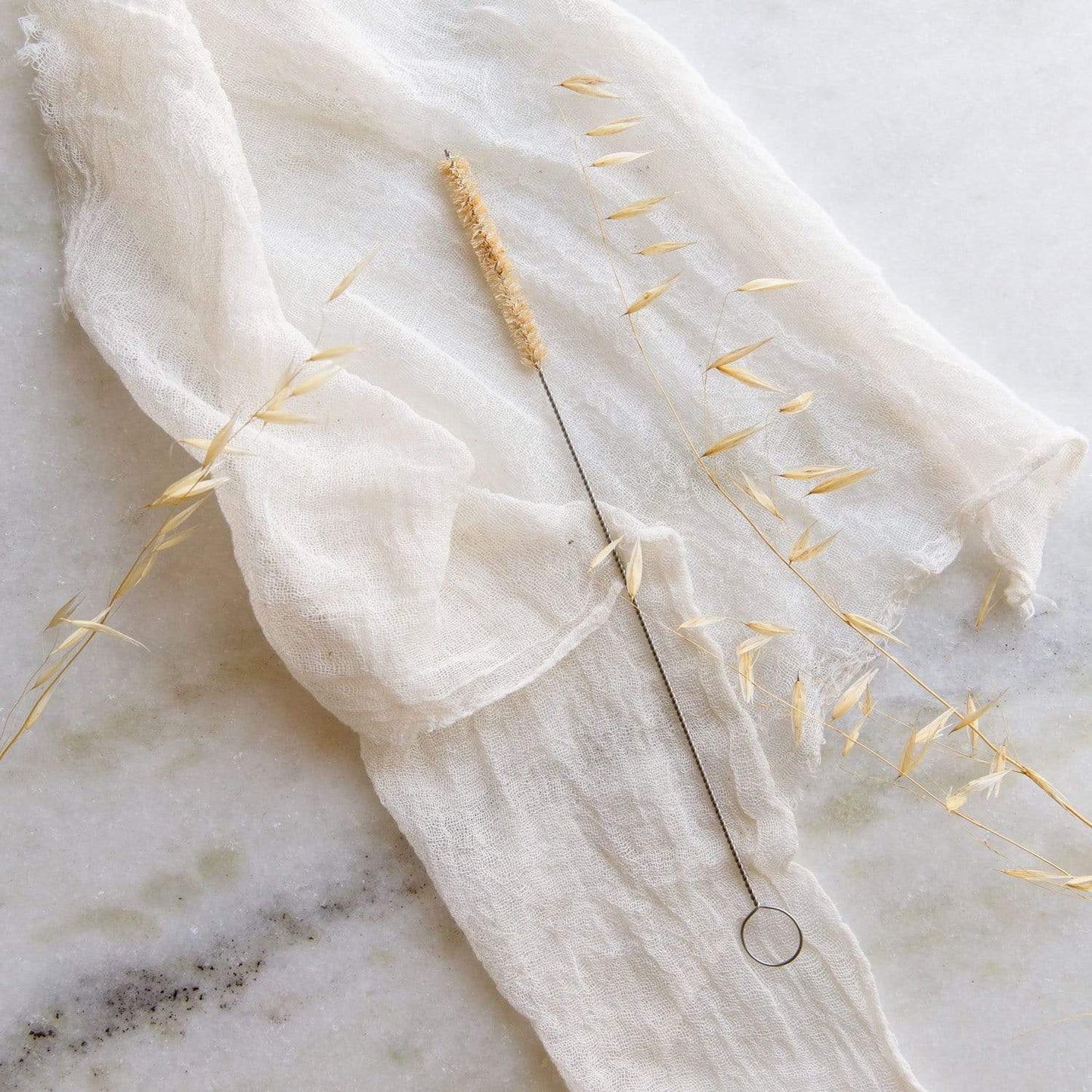 The Straw Cleaner from ZeroWasteStore.com, featuring natural bristles and a metal handle, is displayed on off-white fabric with dried oat stems on a light marble surface.