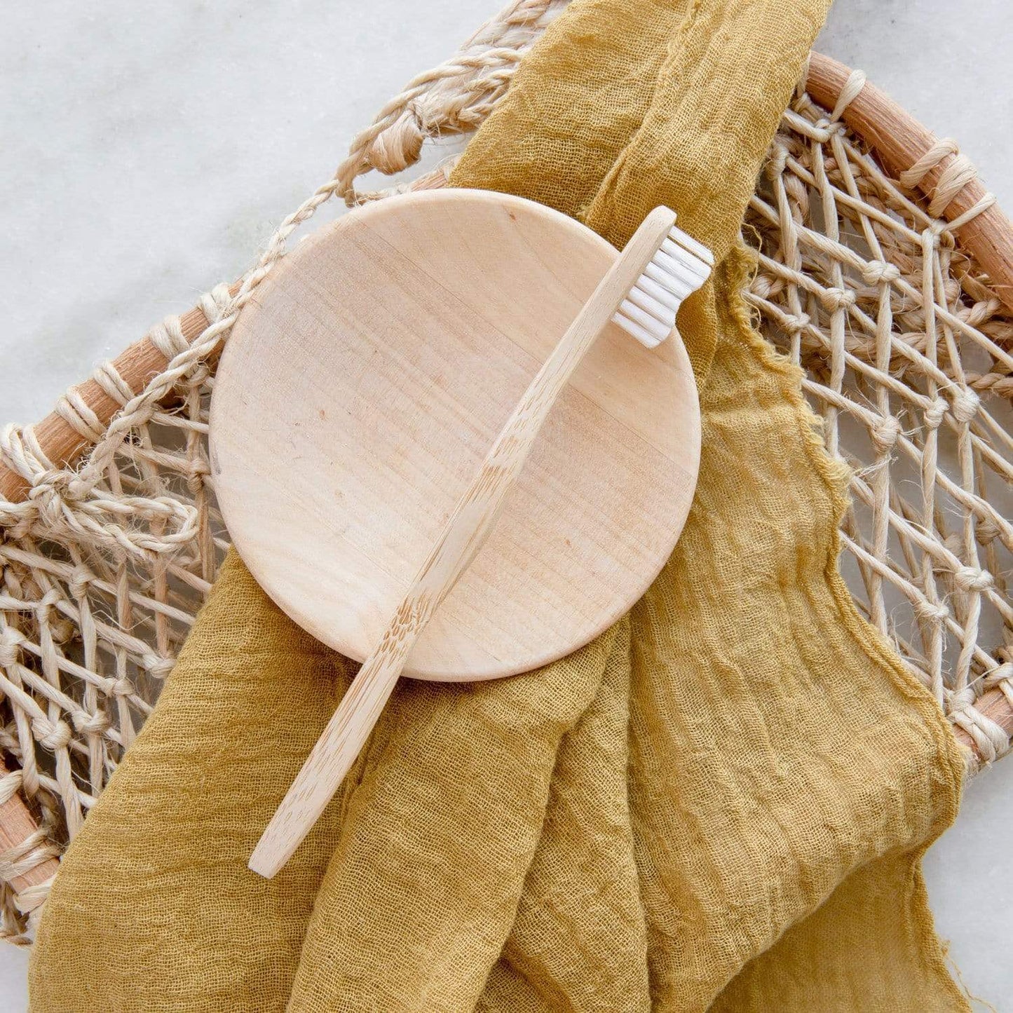 A Bamboo Toothbrush - Child by ZeroWasteStore.com rests on a round wooden soap dish atop a woven basket, with a mustard yellow cloth draped beneath.