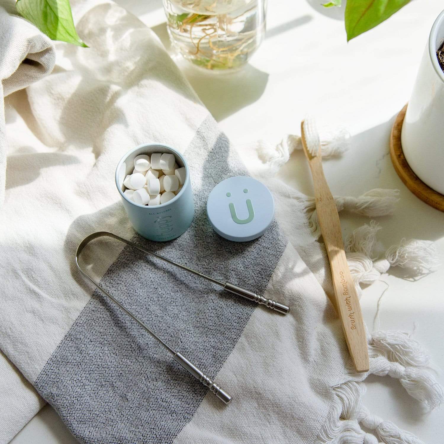 A ZeroWasteStore.com Tongue Scraper—100% stainless steel—sits with chewable toothpaste tablets and a bamboo toothbrush on a striped cloth near a potted plant, creating a chic zero waste setup in bright sunlight.