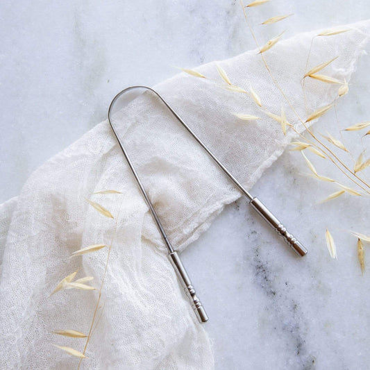 The ZeroWasteStore.com Tongue Scraper—crafted from 100% stainless steel—is a zero waste oral care essential, shown on a white cloth with dried grass atop marble for an eco-friendly touch.