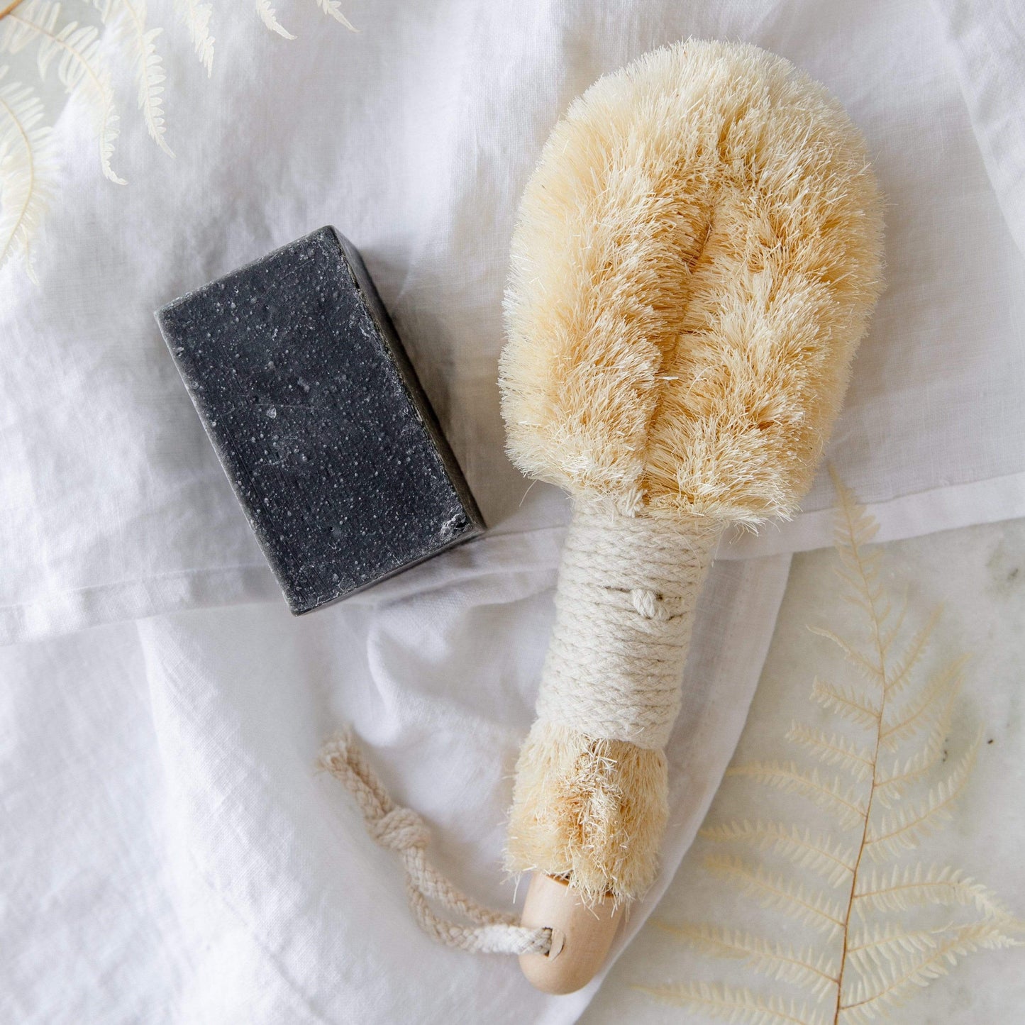The ZeroWasteStore.com Dry Body Brush with sisal bristles and wooden handle sits next to a black rectangular soap bar on white fabric with light fern leaves—ideal for lymphatic drainage rituals.