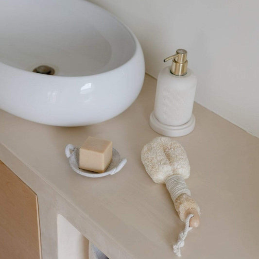 A minimalist bathroom counter displays a white soap dish, a bar of soap, and the Dry Body Brush by ZeroWasteStore.com with sisal bristles and rope handle, alongside a white soap dispenser near a modern sink.