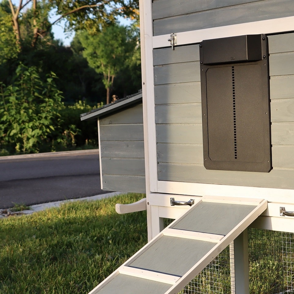 The My Pet Chicken Farmight Automatic Chicken Coop Door (Aluminum) is shown installed on a modern gray coop with black door, small entrance ramp, and greenery and a road in the background for added predator protection.